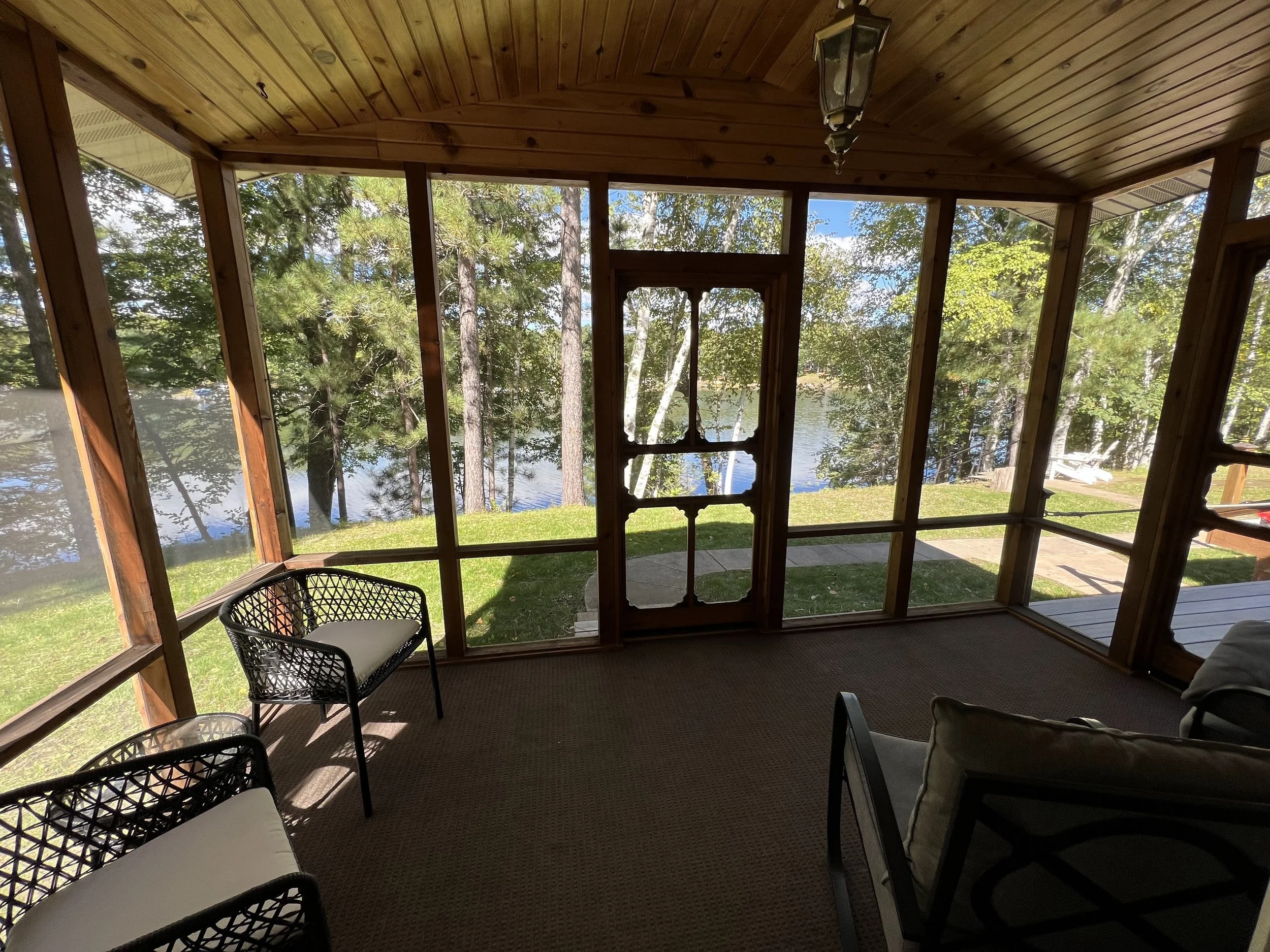 Sunroom with wooden ceiling, screened windows, lake view, chairs, and a bench, overlooking a grassy yard and trees near the lake.