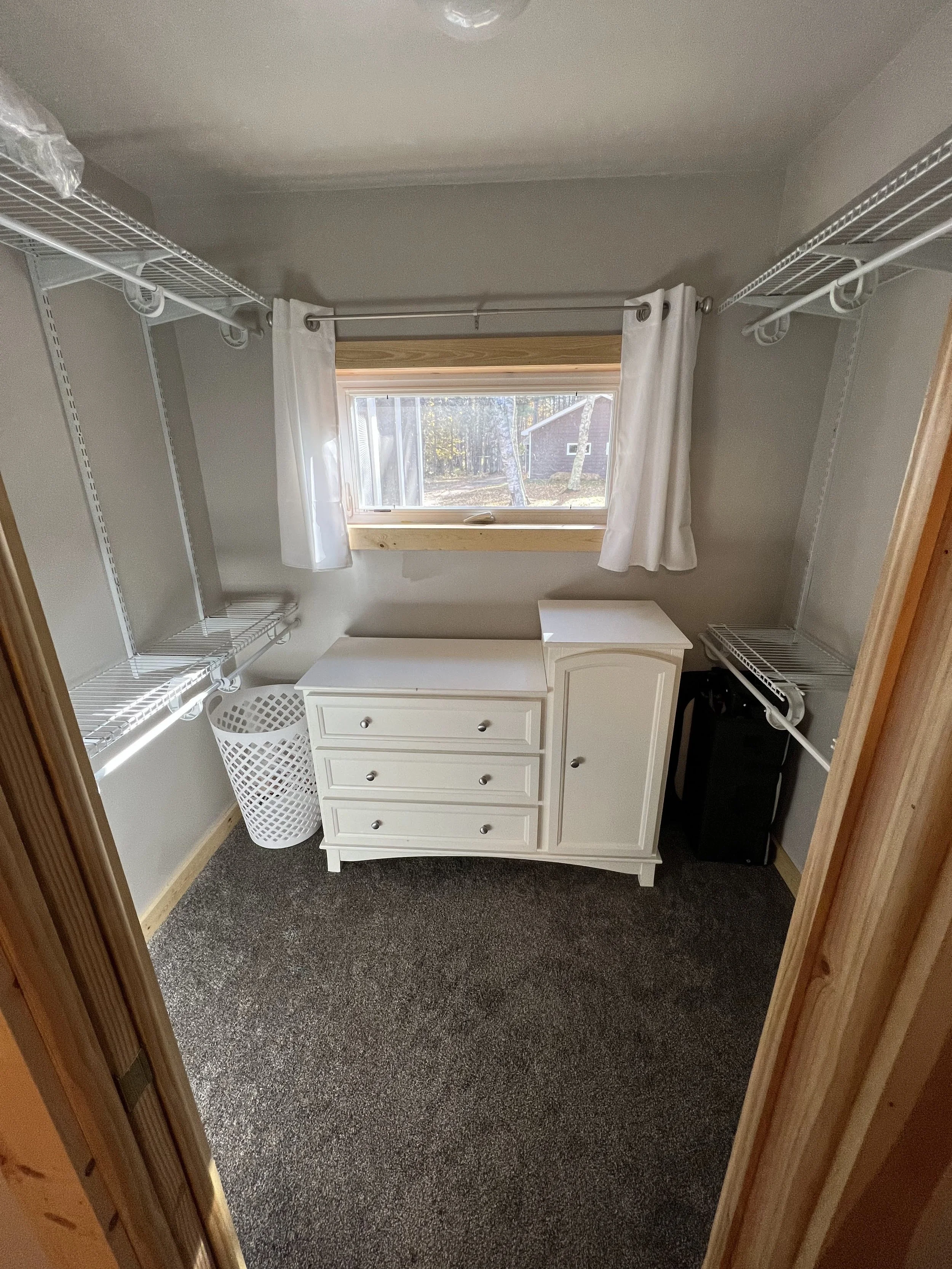 A small closet with white wire shelves on both sides, a window with white curtains, a white dresser, and a laundry hamper inside.