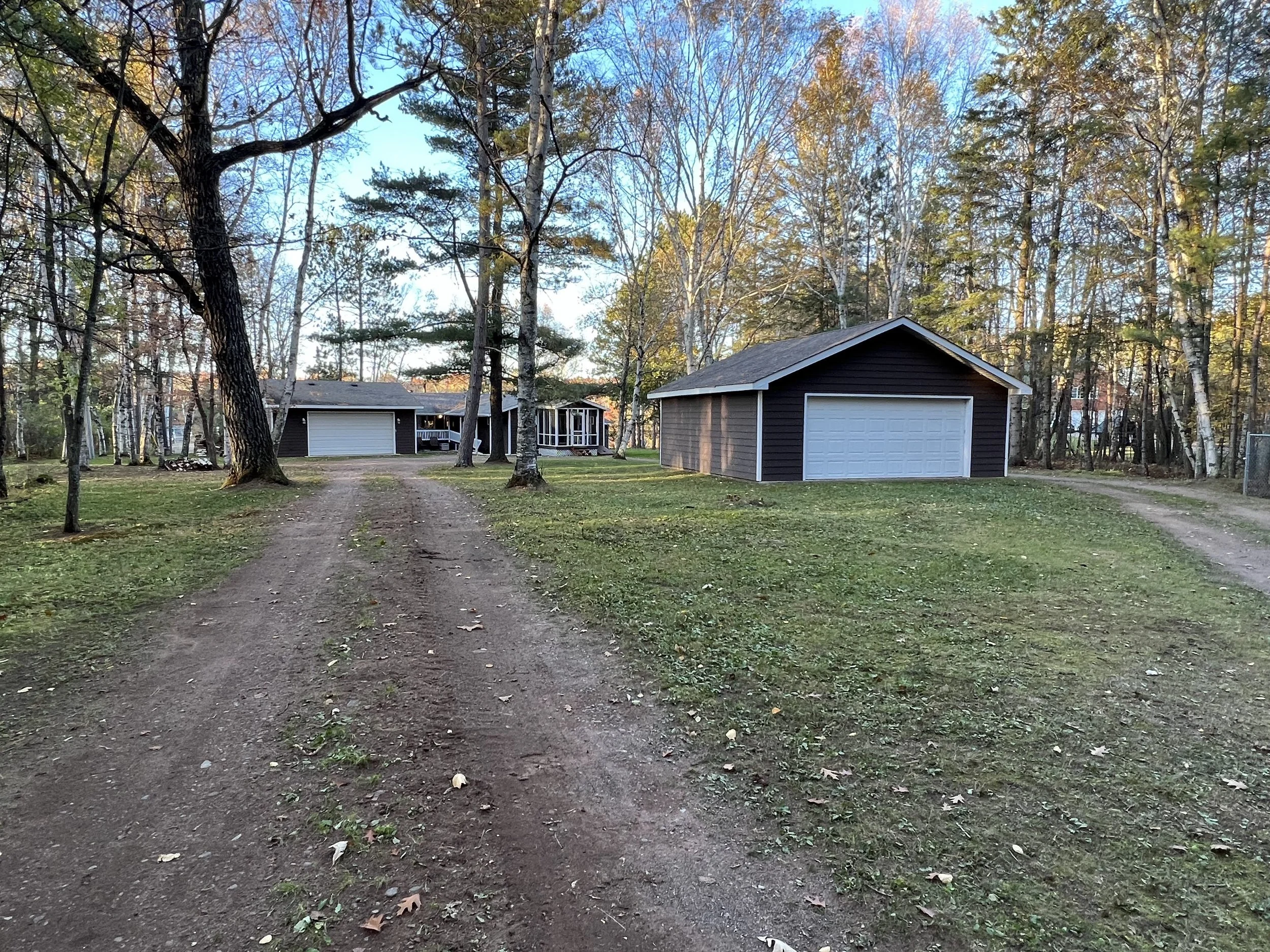 A dirt driveway leads to a house with a two-car garage and a separate garage in a wooded yard with trees and grass