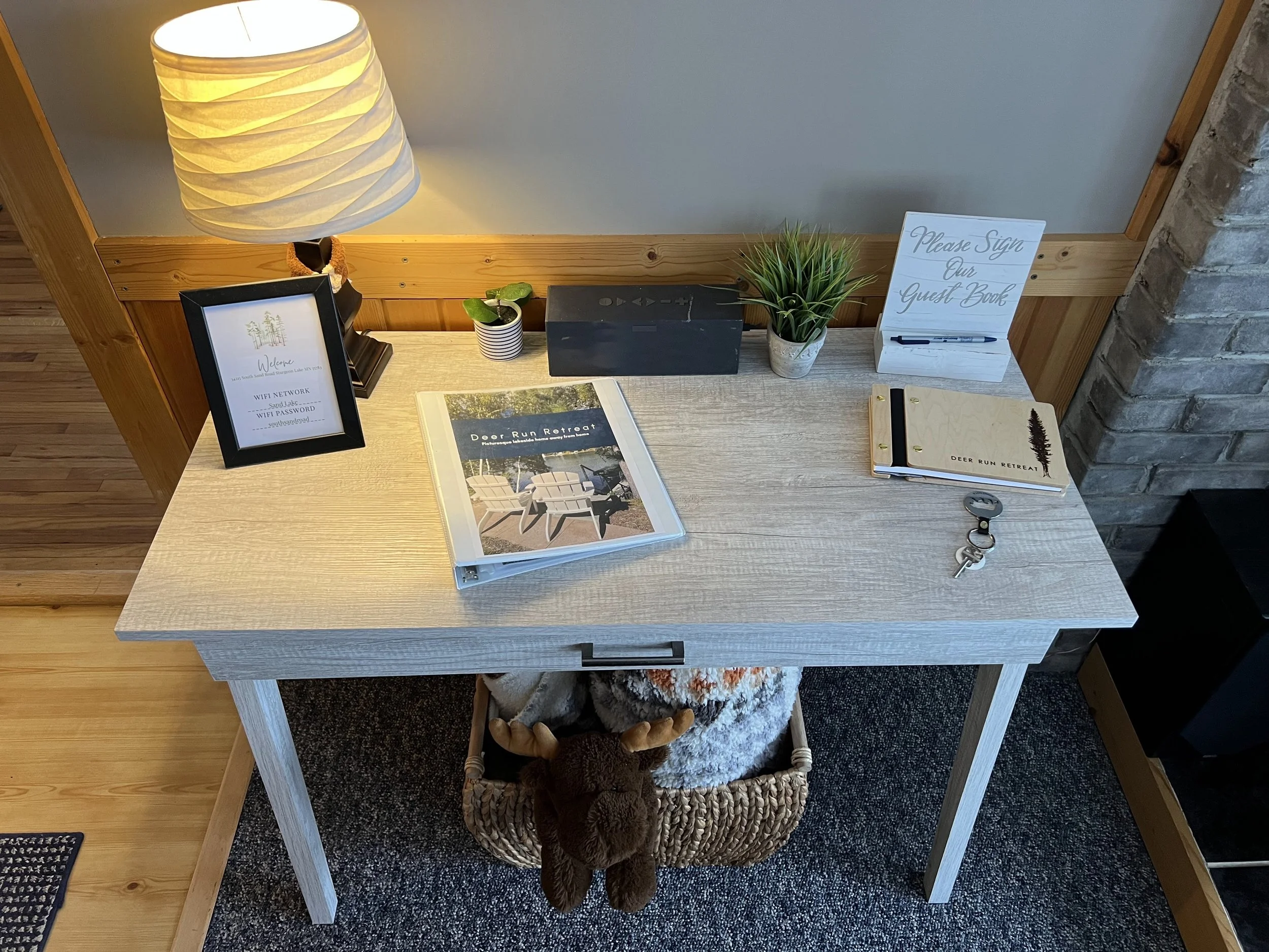 A desk with a lamp, framed welcome sign, potted plants, a black box, guest book, keys, and a stuffed reindeer in a basket underneath.