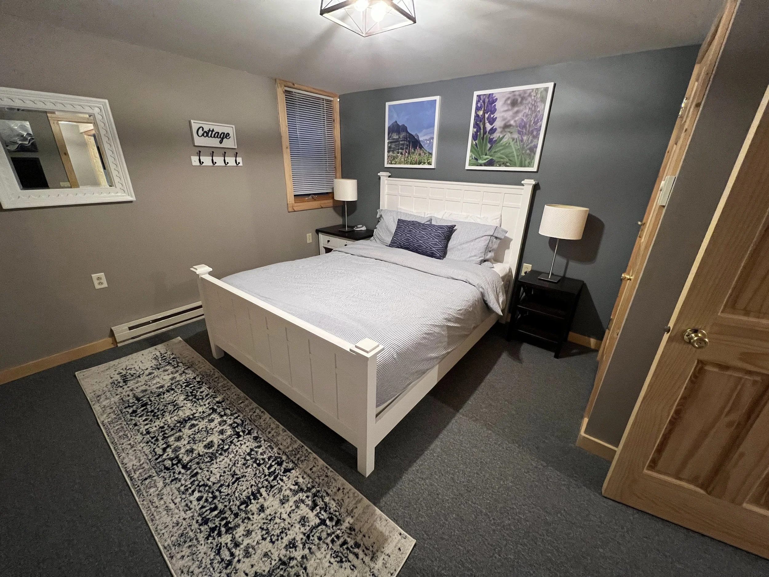 A cozy bedroom with a white bed, gray bedding, and decorative pillows. There are black nightstands with lamps on each side of the bed, a gray accent wall with two framed pictures of mountains and purple flowers, a window with blinds, a mirror on the 