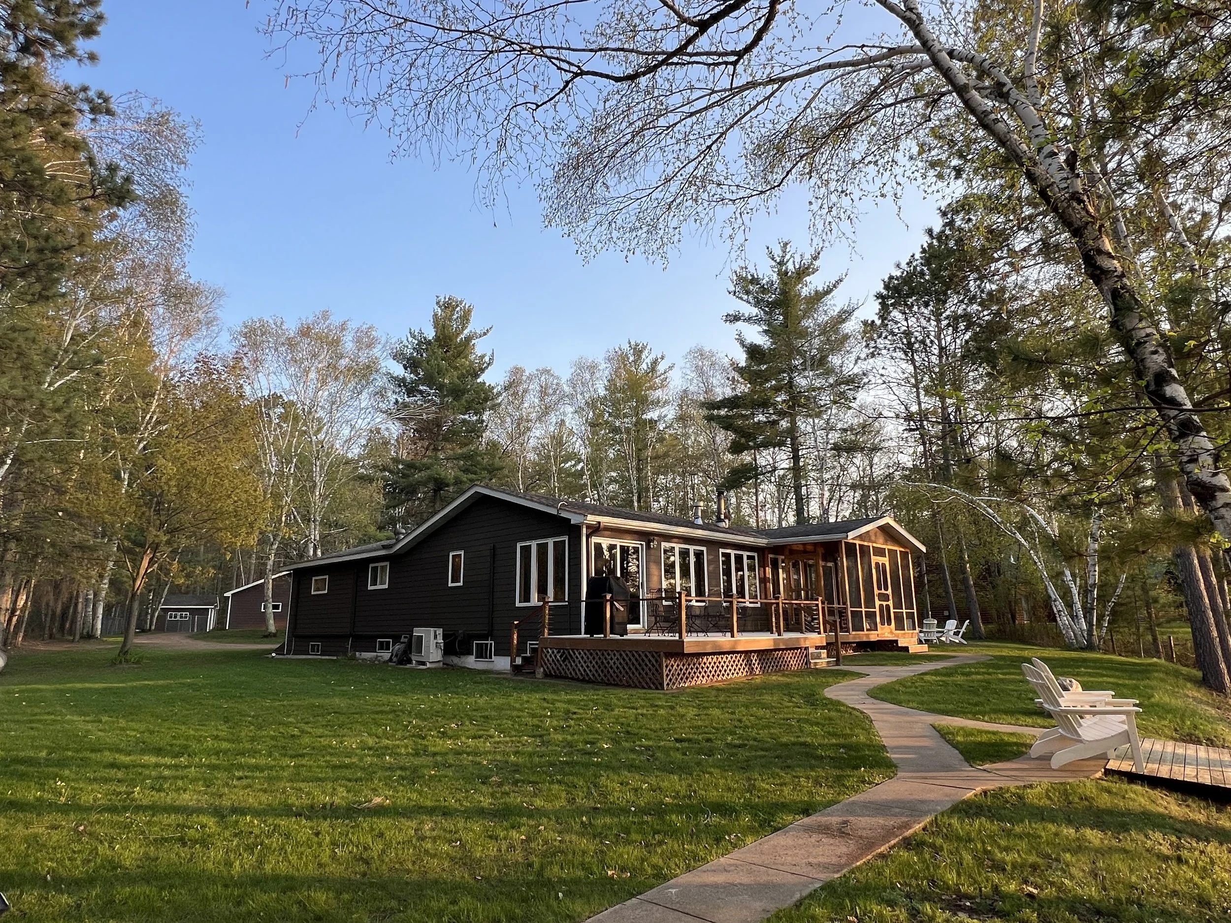 A black house with a wooden deck, surrounded by a green lawn and trees, under a clear blue sky.