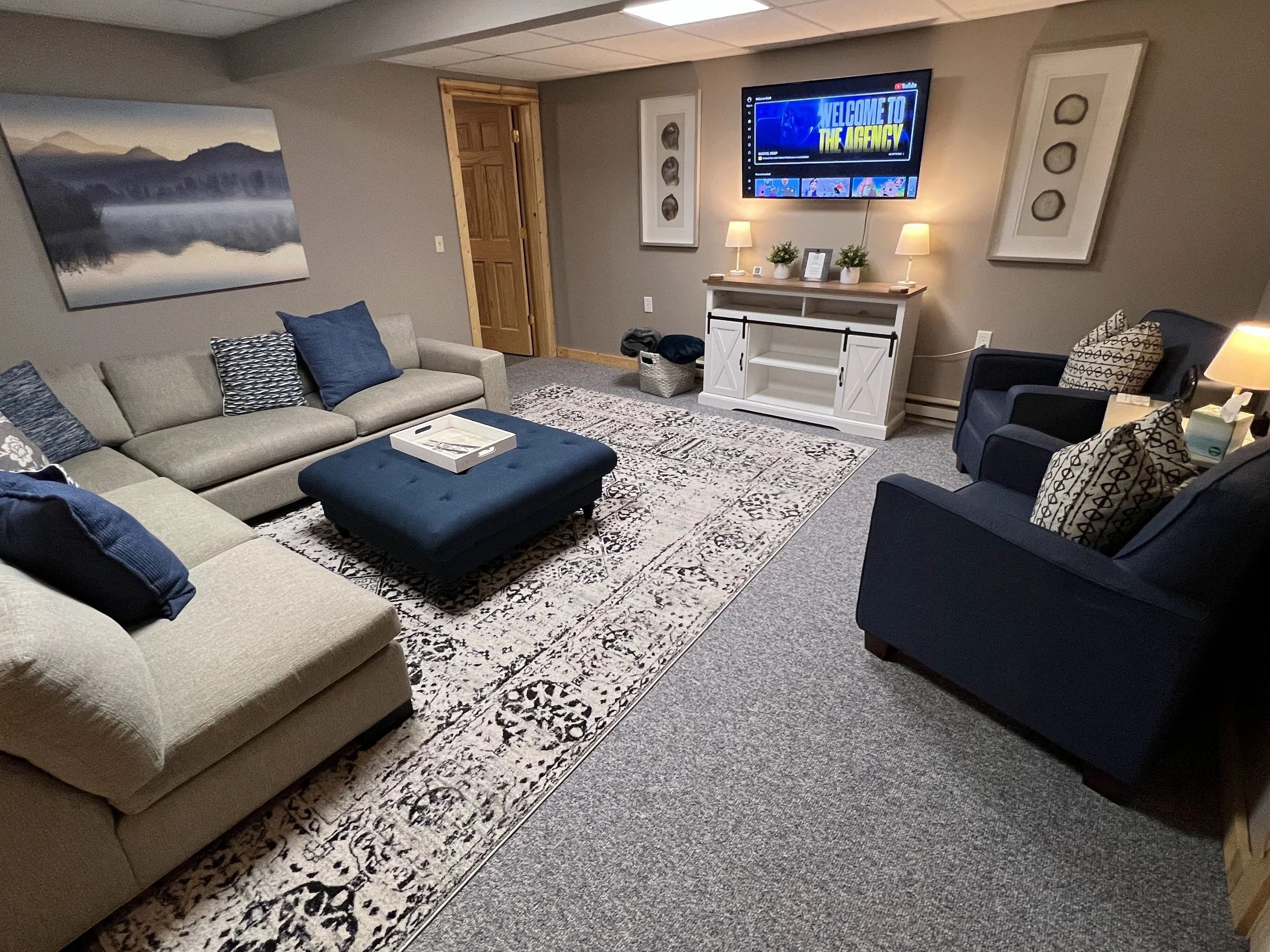 Living room with beige and navy blue sofas, a patterned area rug, a wall-mounted TV displaying 'Welcome to the Agency,' framed wall art, a white media console, side lamps, and decorative pillows.