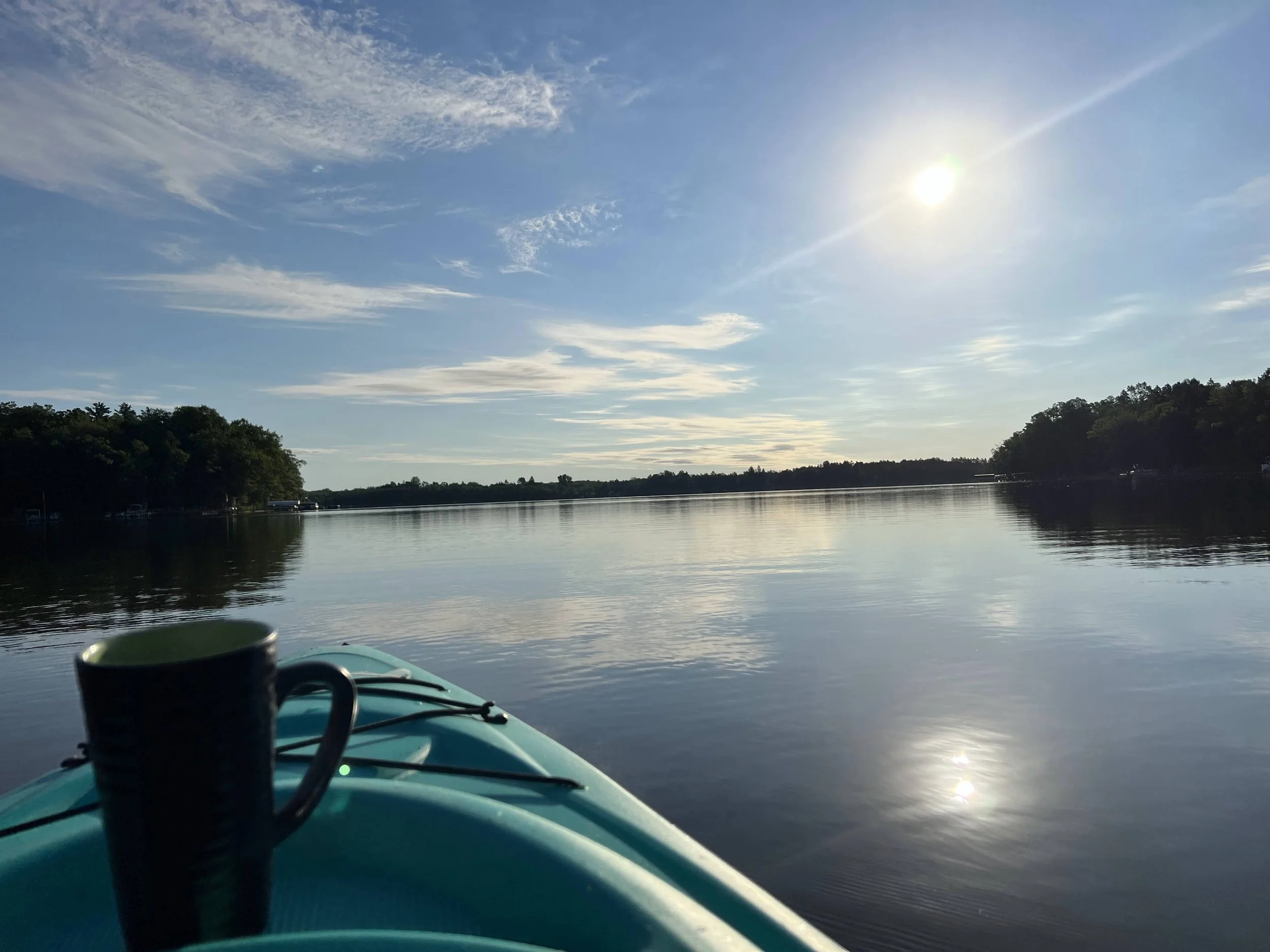 View from a kayak on a calm river with the sun shining in a mostly clear sky and trees lining the horizon.