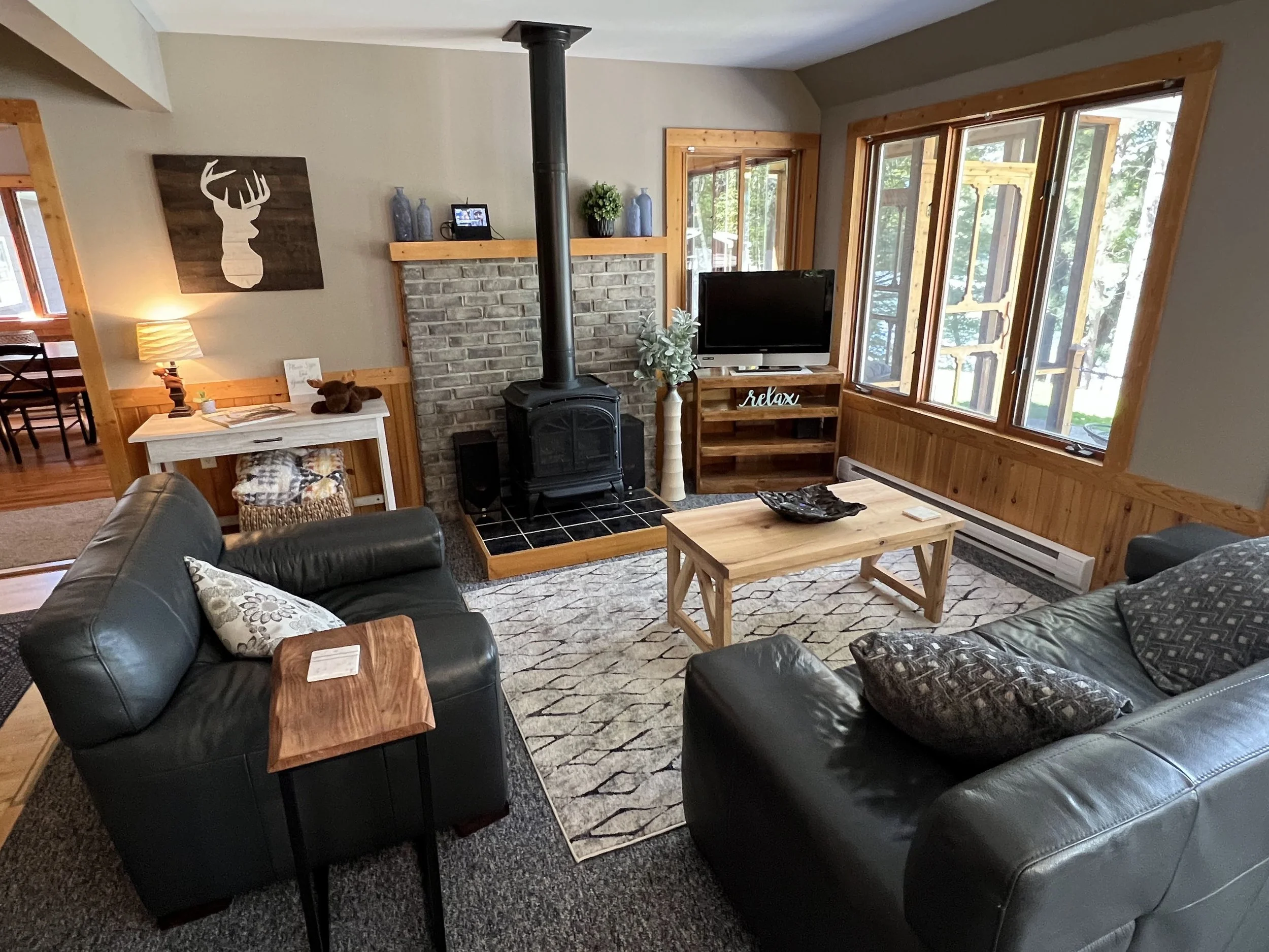 Living room with black leather sofas, wooden coffee table, fireplace, and large windows showing trees outside.