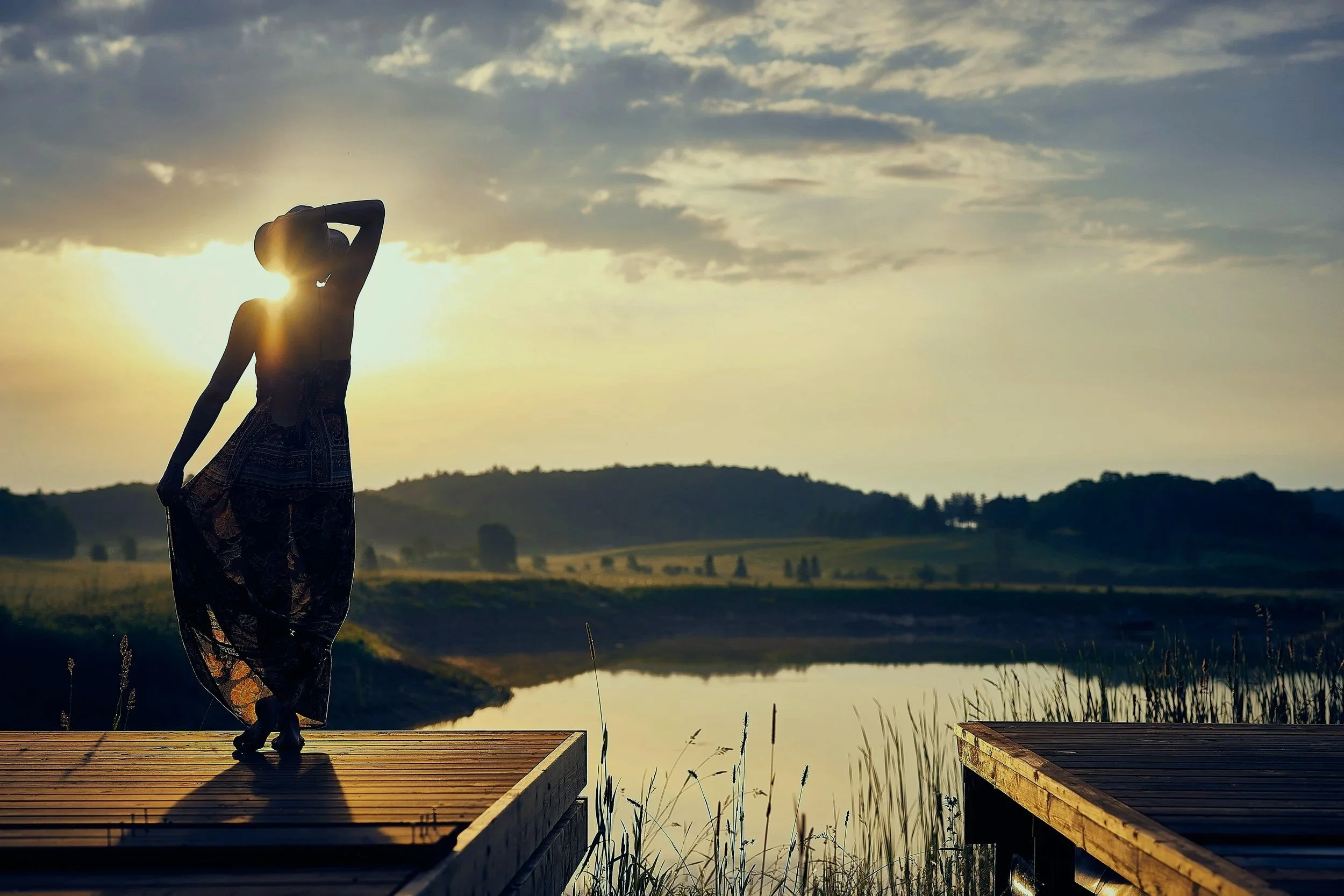 Silhouette of a woman wearing a hat and a flowing skirt standing on a wooden dock by a lake at sunset.