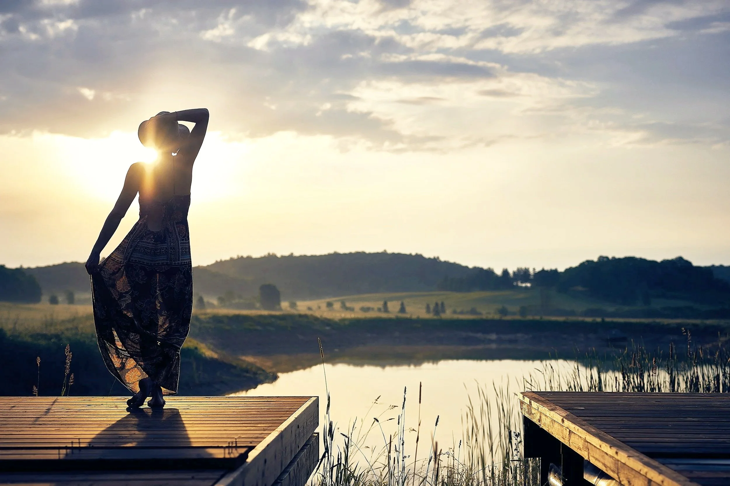 A silhouette of a woman standing on a wooden dock beside a lake during sunset, with hills and a partly cloudy sky in the background.