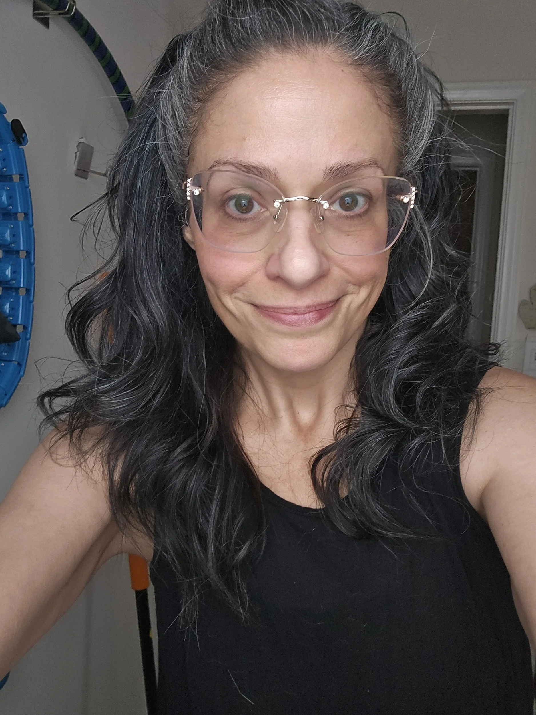 Close-up selfie of a woman with curly black and gray hair, wearing glasses, smiling, and dressed in a black sleeveless top.