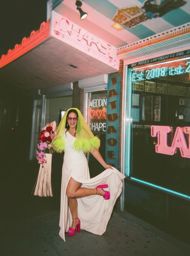 BRIDE wearing a white WEDDING dress with pink high heels and pink sunglasses holding a bouquet of flowers outside a tattoo shop at night.