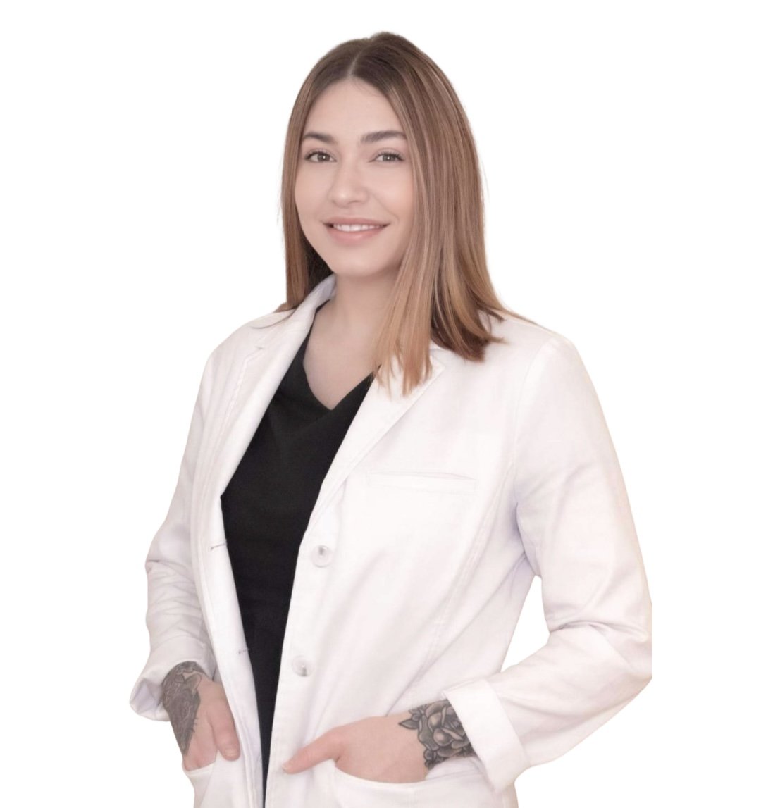 Young woman with light skin, shoulder-length light brown hair, wearing a white jacket over a black top, standing against a plain white background.