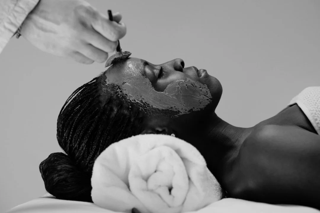 A woman receiving a facial treatment, lying on a spa bed with her eyes closed while a practitioner applies a facial mask. A rolled towel is beneath her head.