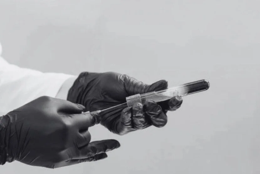 Close-up of a person wearing black gloves holding a blood test tube with a needle and syringe, preparing for a medical blood draw.