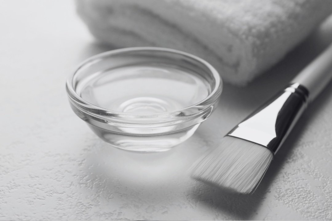 A small glass bowl filled with clear water, a paintbrush with a silver handle, and a folded towel in the background on a white textured surface.