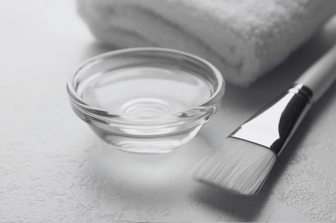 A clear glass bowl filled with water, a folded towel, and a paintbrush with a metallic ferrule on a white textured surface.