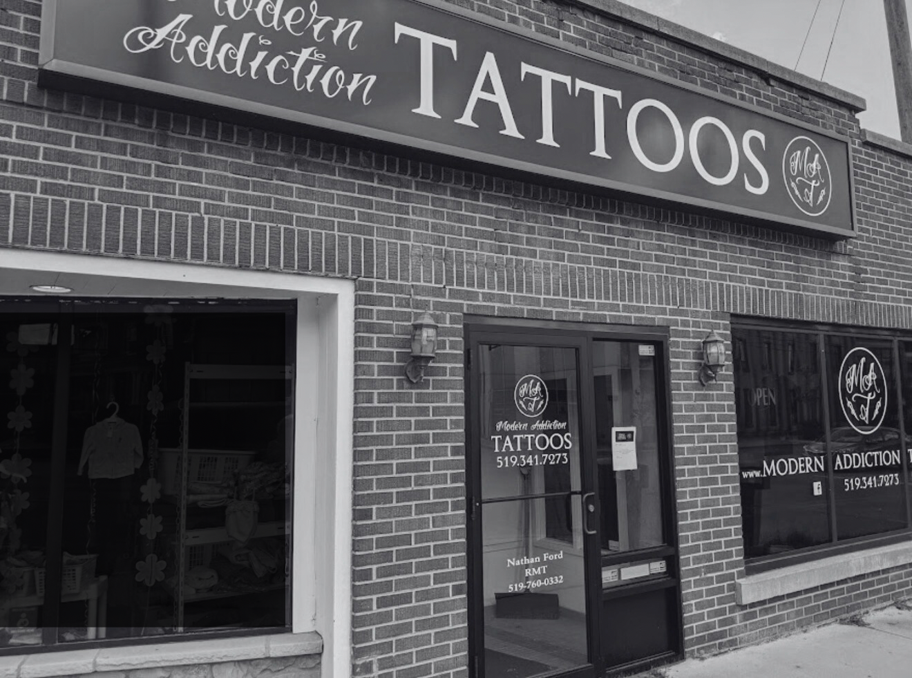 Exterior of a brick storefront with a sign reading 'Modern Addiction TATTOOS' and additional signage on the door and window including contact information.