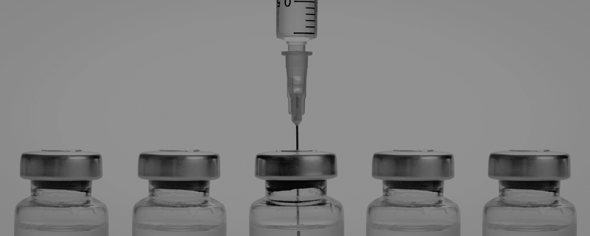 A close-up of a syringe injecting a liquid into a row of glass vaccine vials with metal caps.
