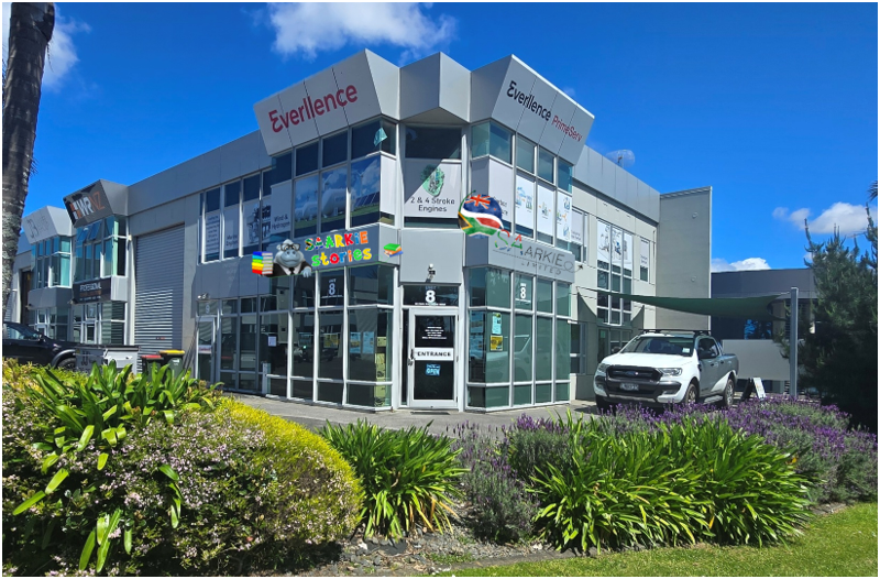 Modern commercial building with glass facade and signage, surrounded by green landscaping and parked cars.