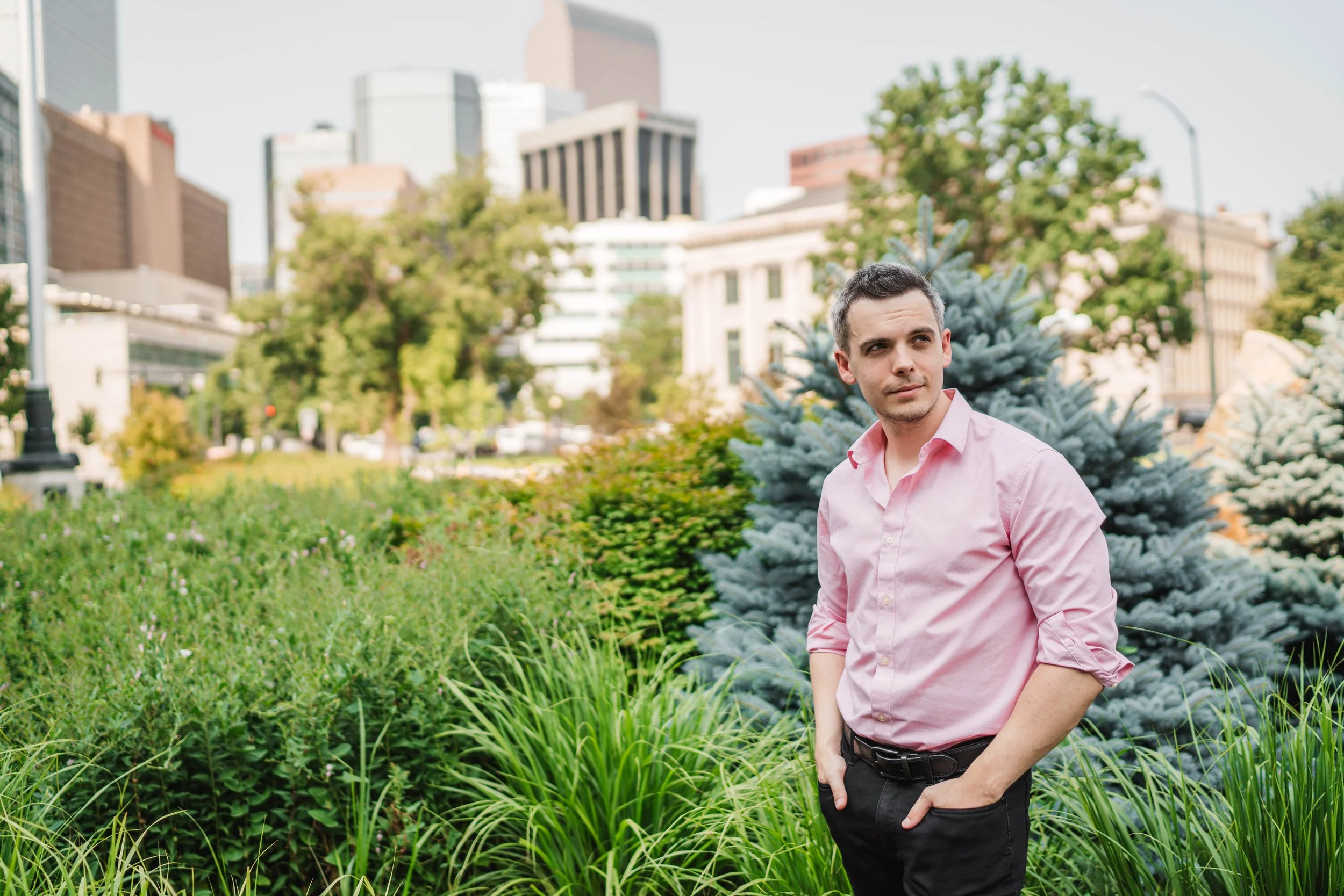 A young man in a pink shirt standing in a city park with green foliage, trees, and buildings in the background.