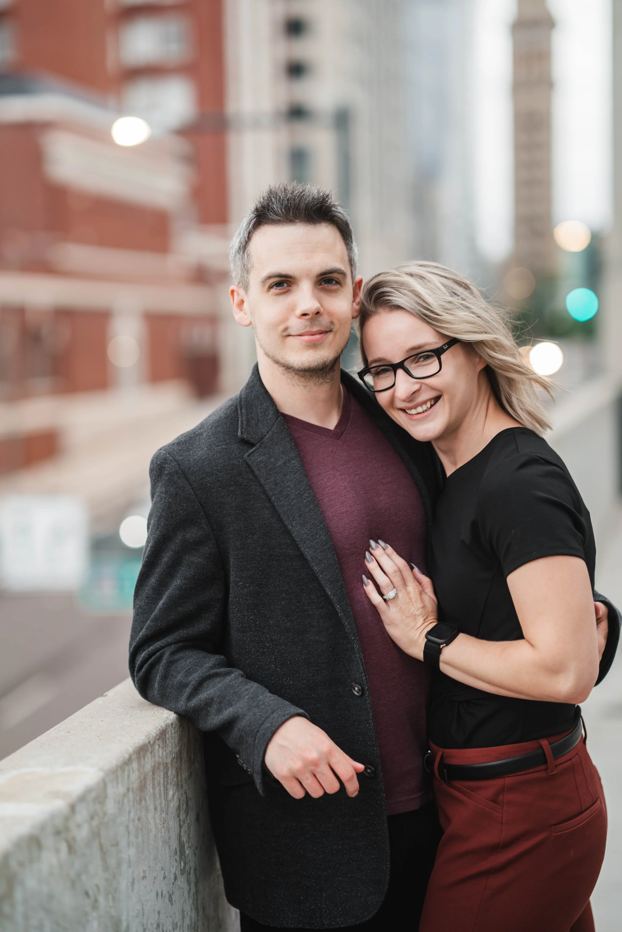 A smiling couple standing on a city sidewalk, embracing each other with buildings and streetlights in the background.