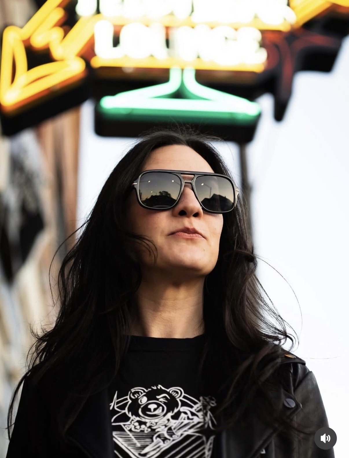 A woman with long dark hair wearing sunglasses and a black leather jacket, standing outside with neon signs in the background.