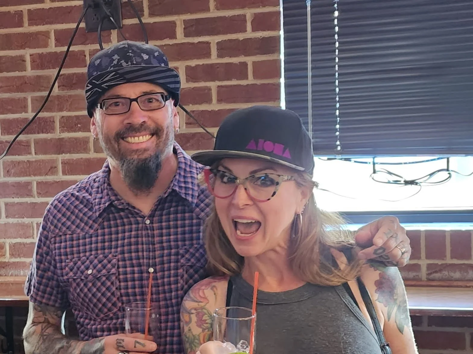 Close-up of a smiling woman with long blonde hair, tattoos on her arm, glasses, and a black cap with purple text, sitting outdoors with a man wearing a striped helmet and red shirt.