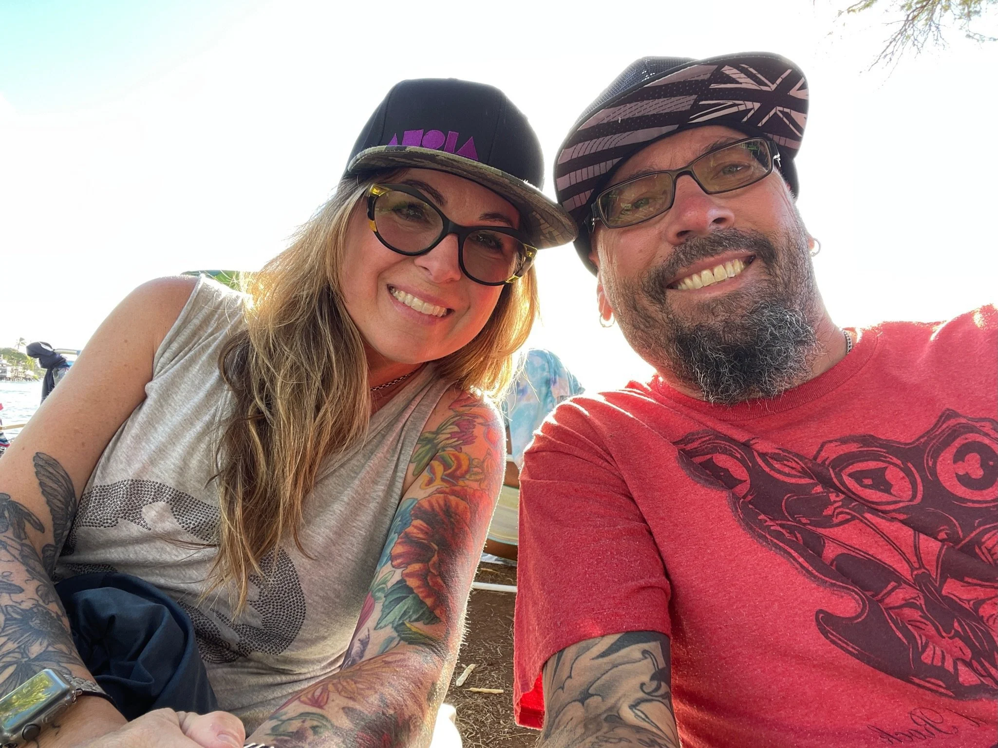 A tattooed bartender wearing glasses, a black baseball cap, and a black T-shirt with a logo, standing behind a bar with bottles and taps, looking at a tablet or menu.