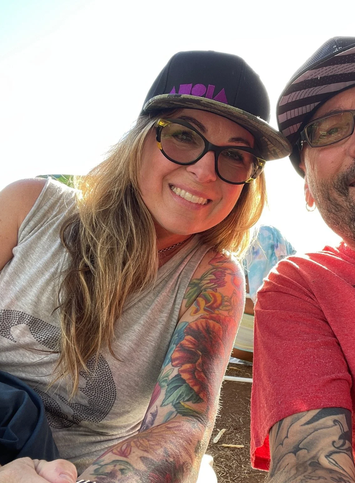 Close-up of a smiling woman with long blonde hair, tattoos on her arm, glasses, and a black cap with purple text, sitting outdoors with a man wearing a striped helmet and red shirt.