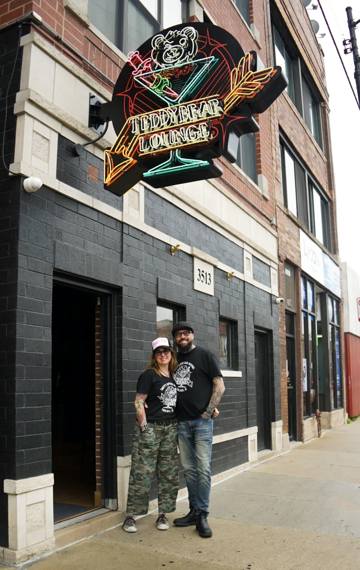 Two people standing outside a black brick building called Teddy Bear Lounge, with a neon sign above them featuring a teddy bear, martini glass, and cocktail umbrella. The woman has tattoos, wears a cap, sunglasses, a black Teddy Bear Lounge T-shirt, and camouflage pants. The man has tattoos, a beard, glasses, a black Teddy Bear Lounge T-shirt, and jeans.