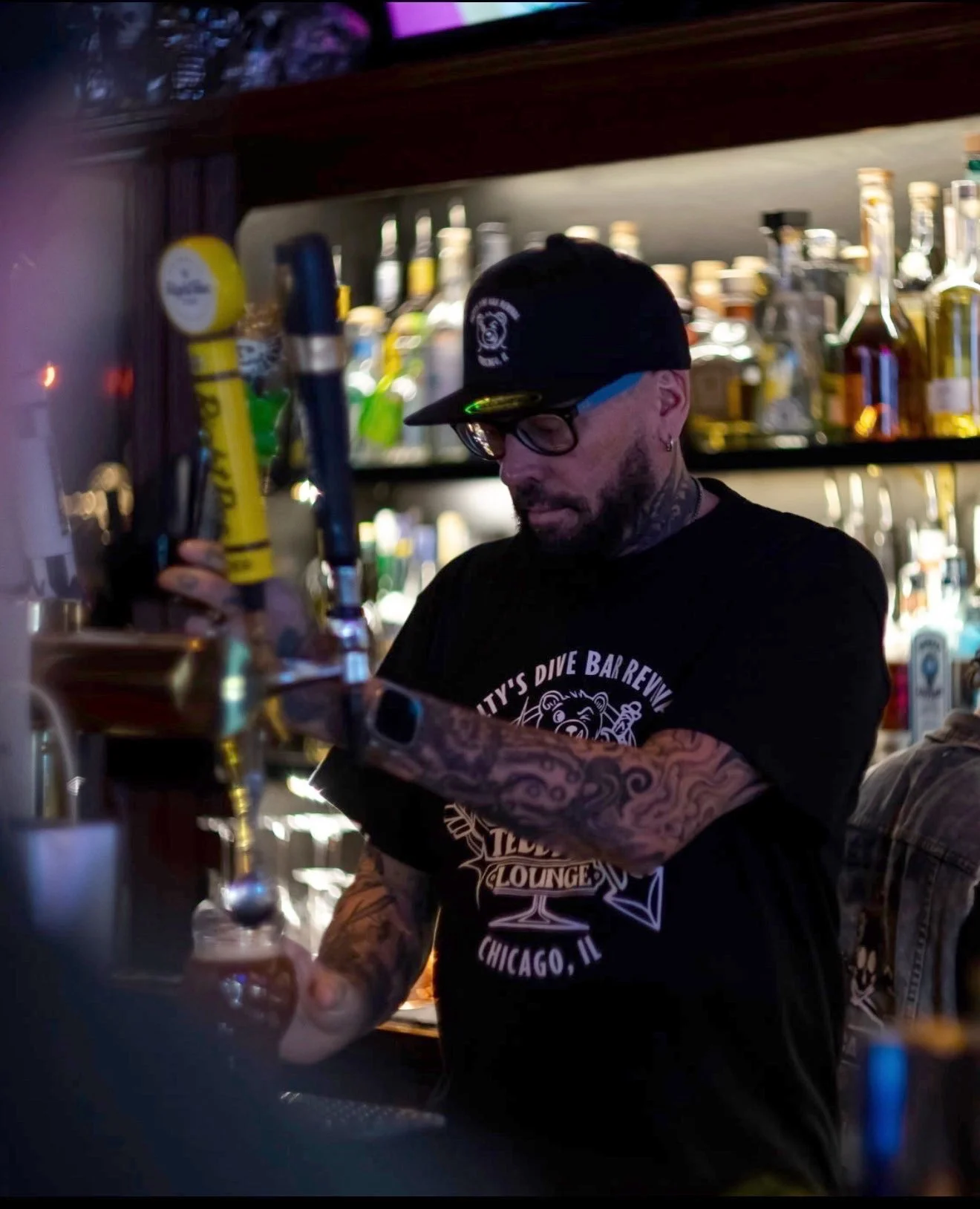 A tattooed man in a black T-shirt, glasses, and a black cap mixes drinks at a bar with liquor bottles behind him.