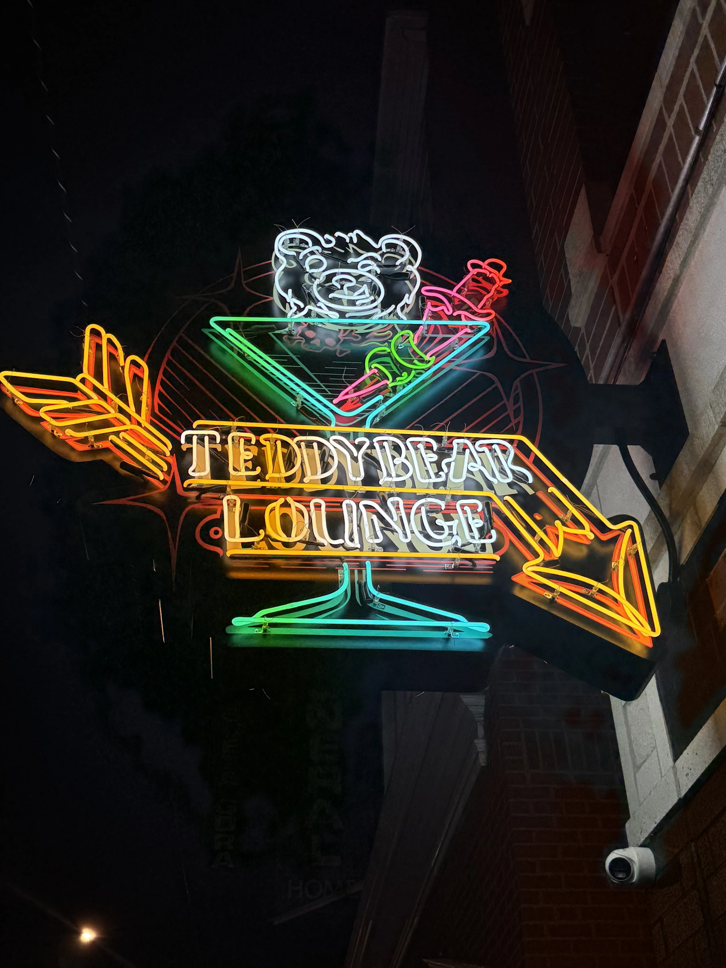 Neon sign for Teddy Bear Lounge featuring a teddy bear head above a cocktail glass with a cherry and lime, with neon arrows and the text 'Teddy Bear Lounge' in lights.