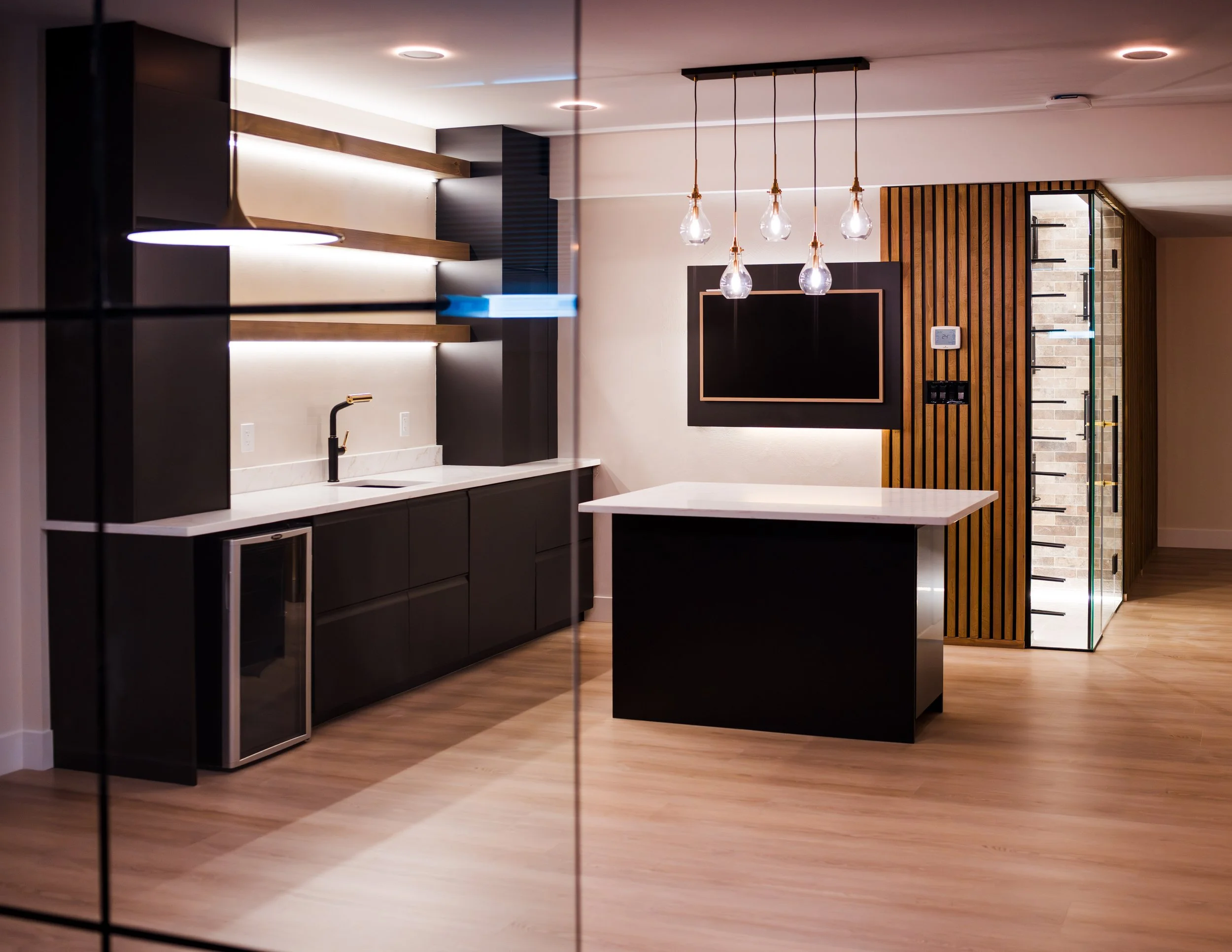 Modern kitchen with black cabinets, a white marble countertop, a black faucet, an island with a white countertop, a wooden slat wall with a mounted blackboard, pendant lights hanging above, and a brick and glass door enclosure.