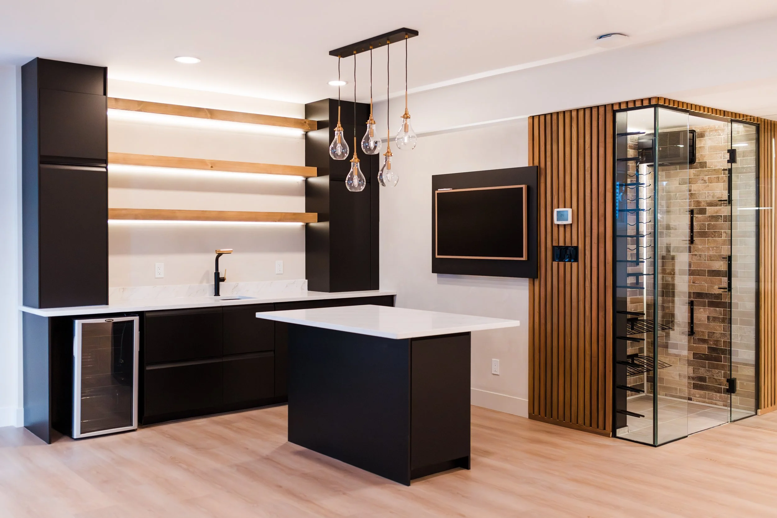 Modern kitchen with black cabinetry, a white marble countertop, a small black refrigerator, a kitchen island, hanging light bulbs, wooden shelves, a wall-mounted TV, and a glass-enclosed wine cellar with brick interior.