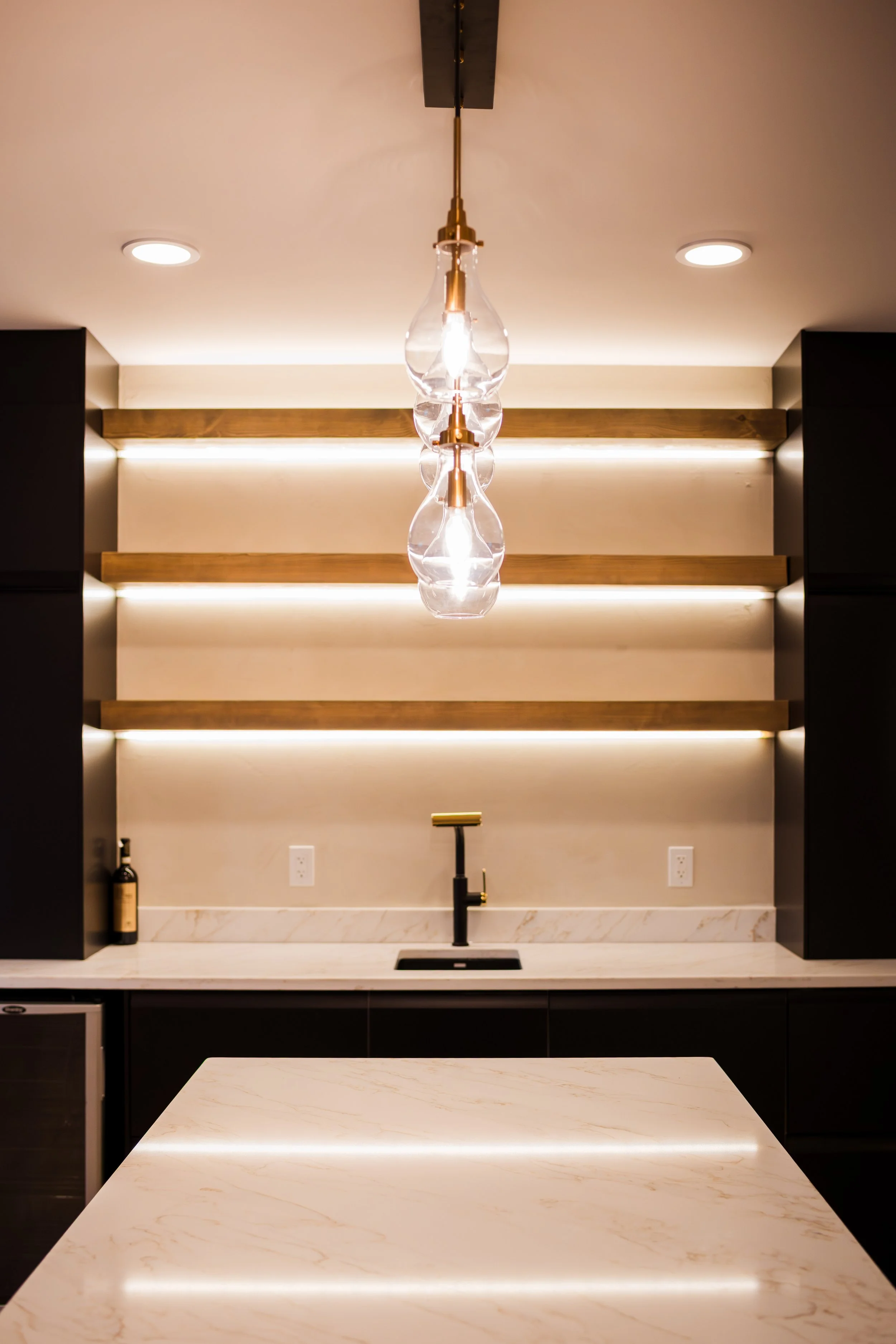 Modern kitchen with a white marble island, black cabinetry, open wooden shelves with LED lighting, black faucet, and a glass pendant light fixture.