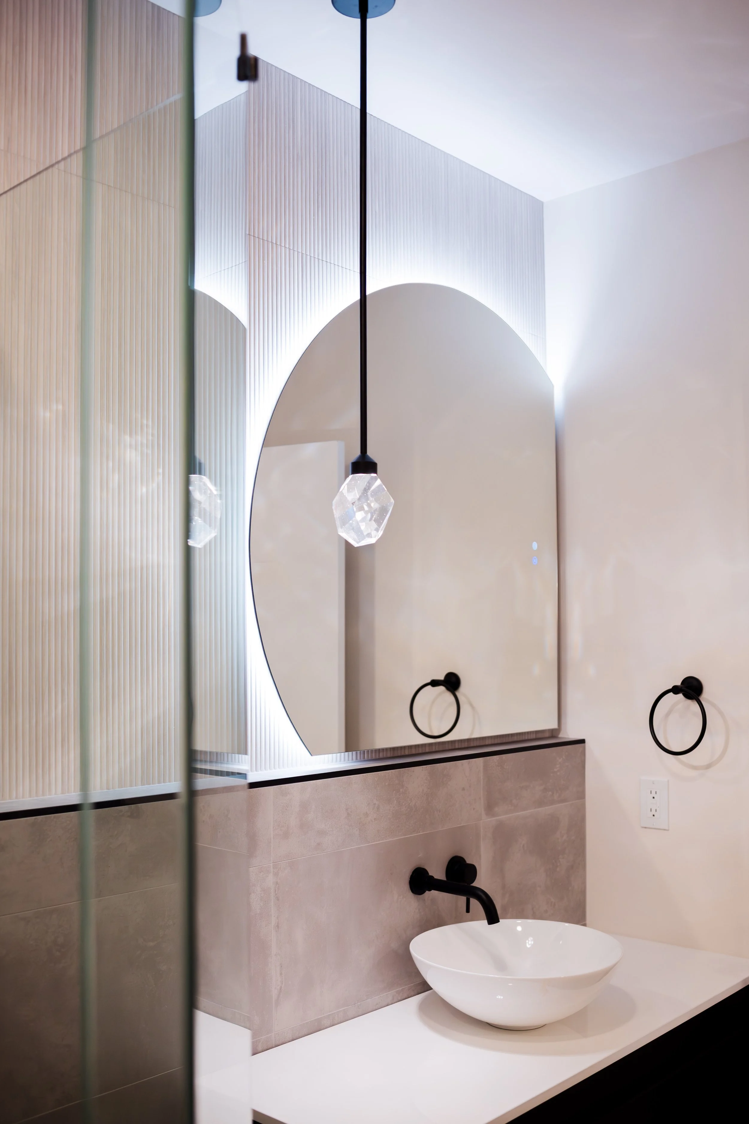 Modern bathroom with a large round mirror, a white vessel sink, and black fixtures. A pendant light hangs from the ceiling, and black towel rings are mounted on the wall.