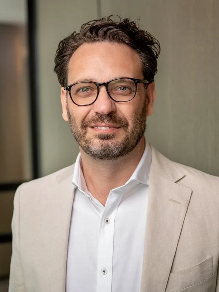 A man with dark hair, beard, and glasses, wearing a cream-colored blazer and white collared shirt, standing indoors.