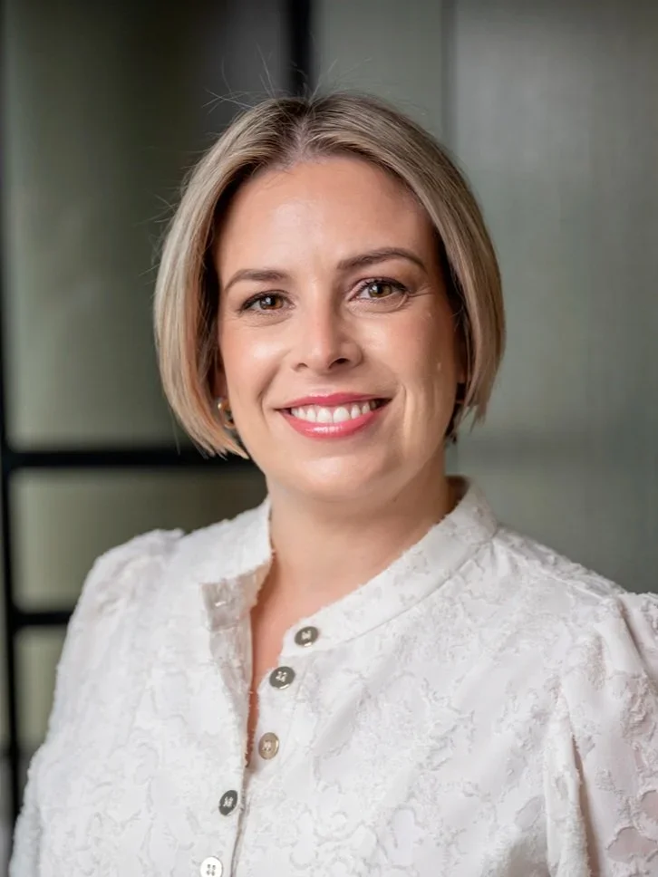 A woman with short blonde hair smiling, wearing a white textured button-up shirt, with a blurred background.