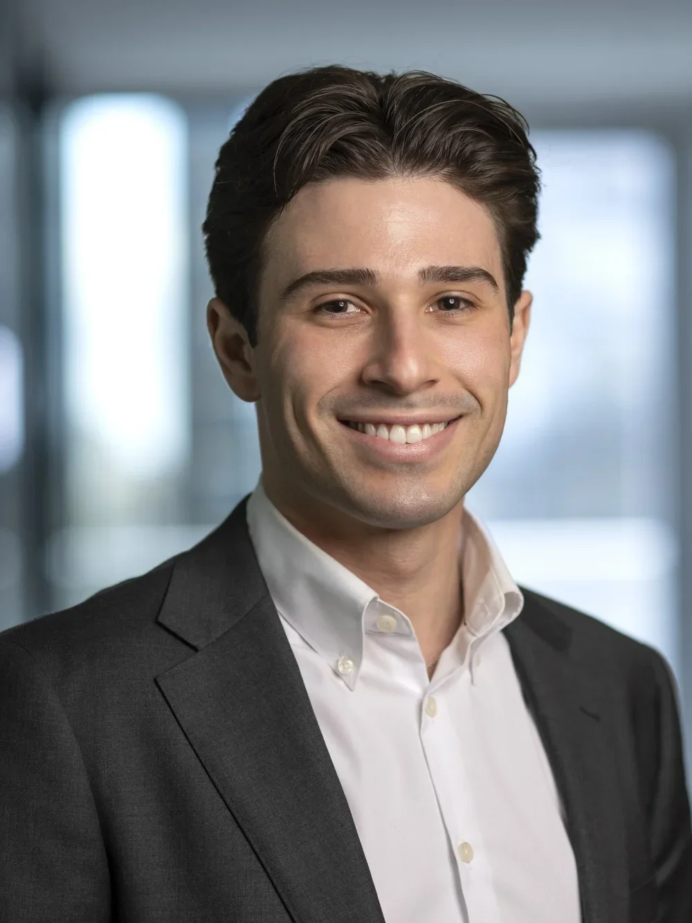 A young man with dark hair and a bright smile, dressed in a white shirt and dark suit, standing in an office setting.