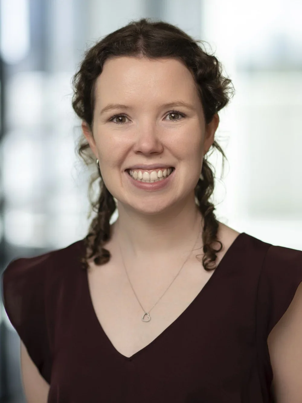 Portrait of a young woman with brown curly hair, smiling, wearing a dark top and a heart-shaped necklace, in a bright indoor setting.