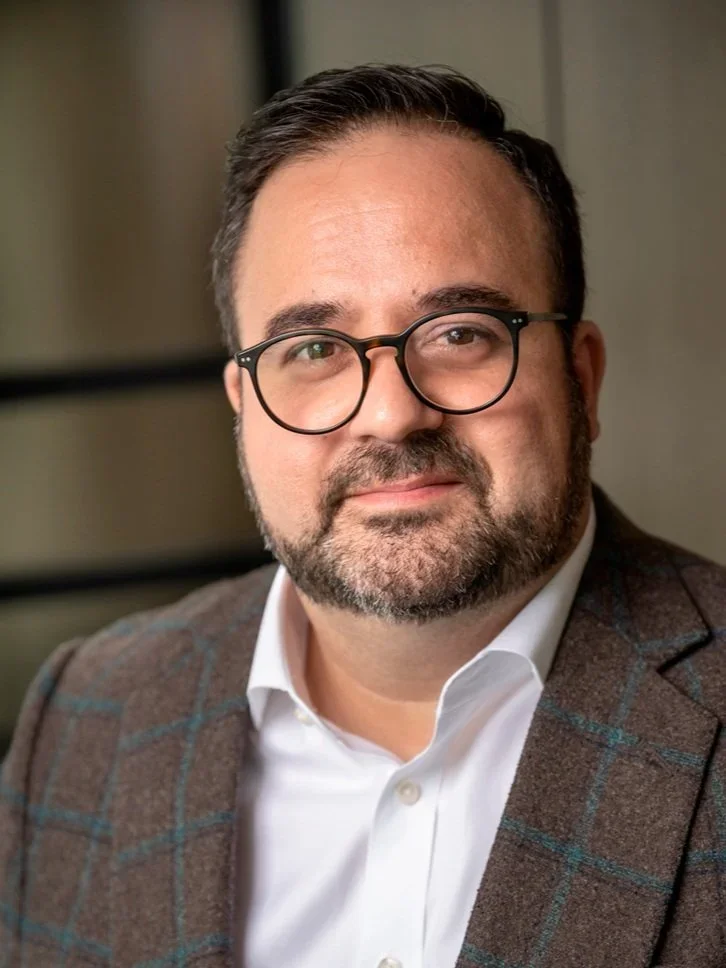 A man with dark hair, glasses, and a beard, wearing a white shirt and a brown checkered blazer, looking at the camera.