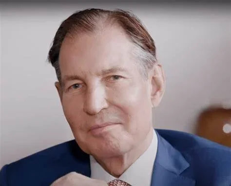 A middle-aged man with brown hair, wearing a dark blue suit and white shirt, smiling slightly at the camera.