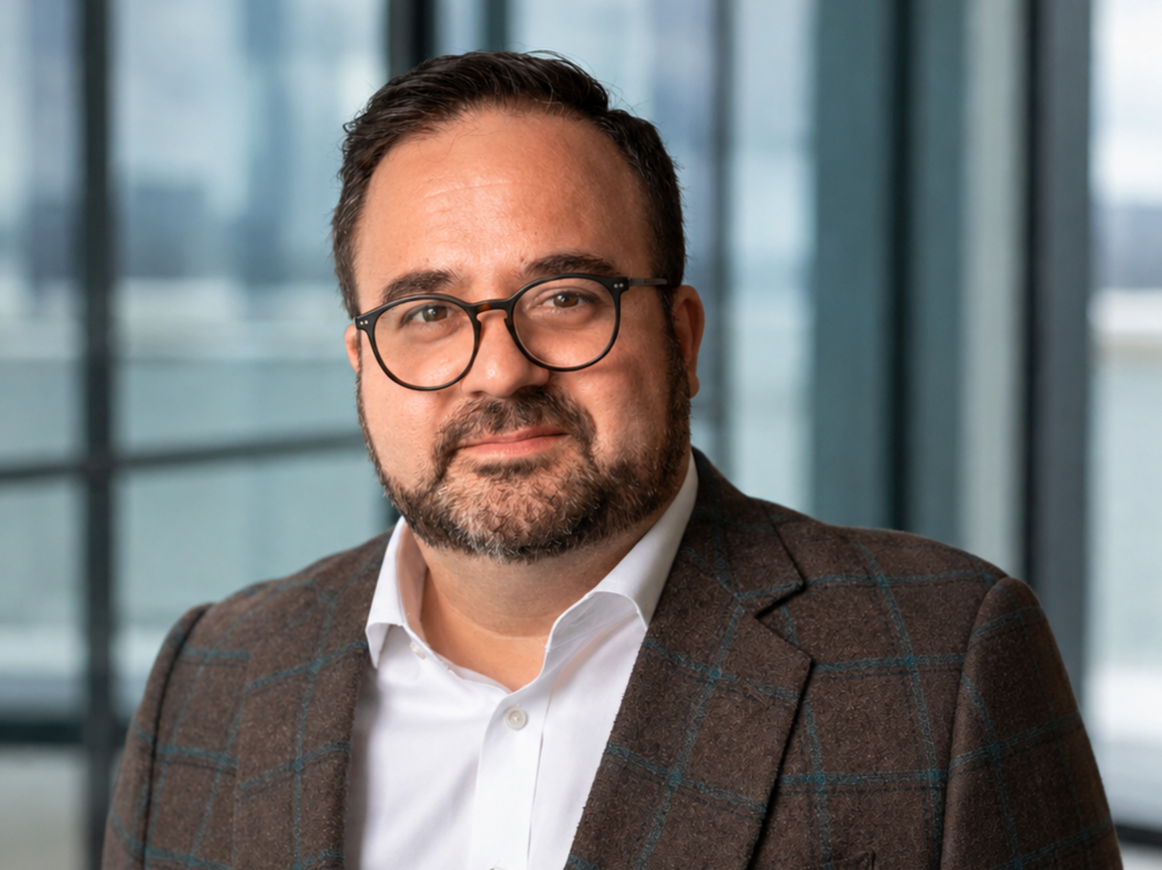 A man with dark hair, glasses, and a beard, wearing a white shirt and a brown checkered blazer, looking at the camera.