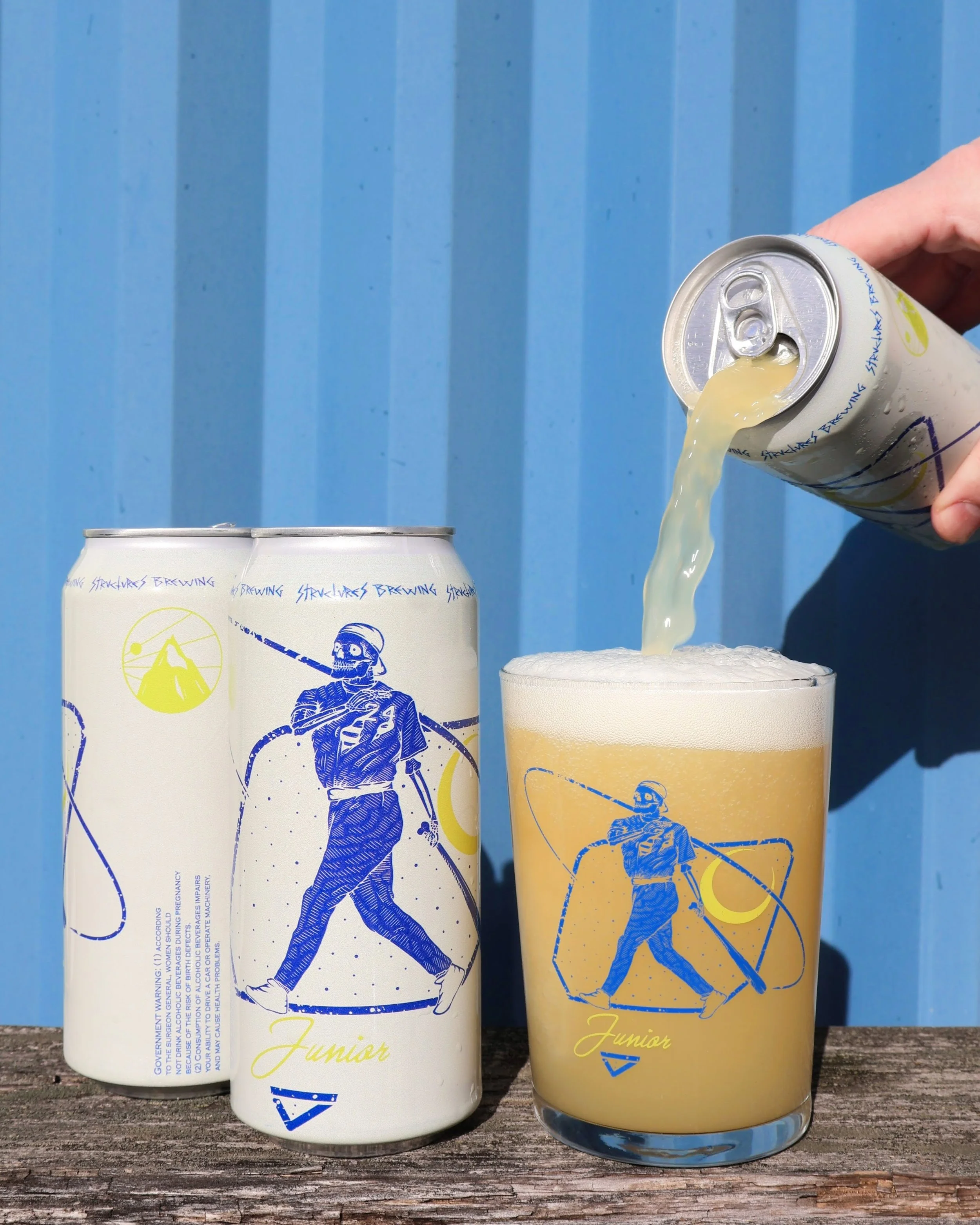 A person pouring a light-colored beverage from a can into a glass with a graphic of a man hiking against a blue background with a wood surface in the foreground.