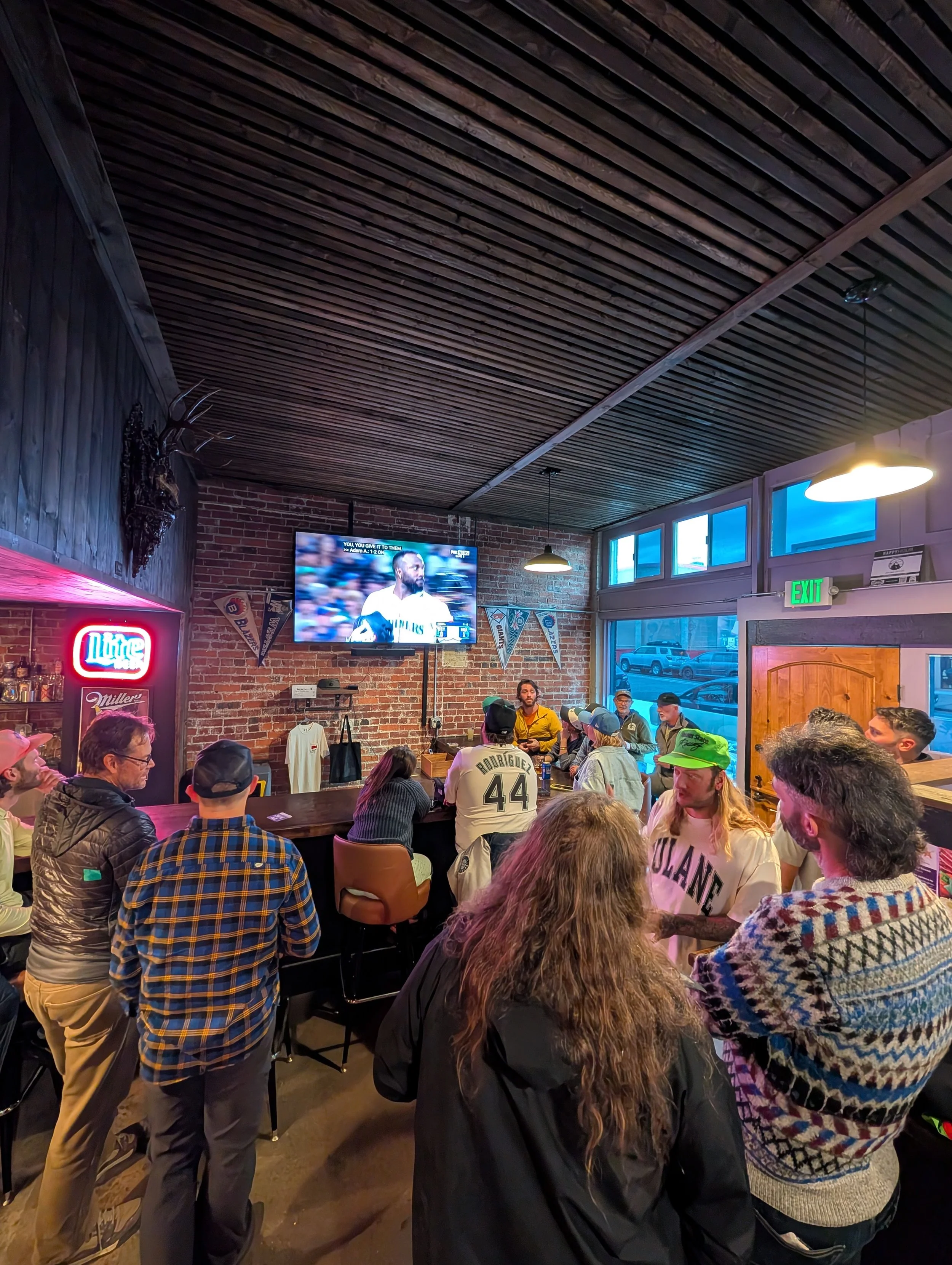 People gathered inside a bar watching a sports game on a television mounted on a brick wall.