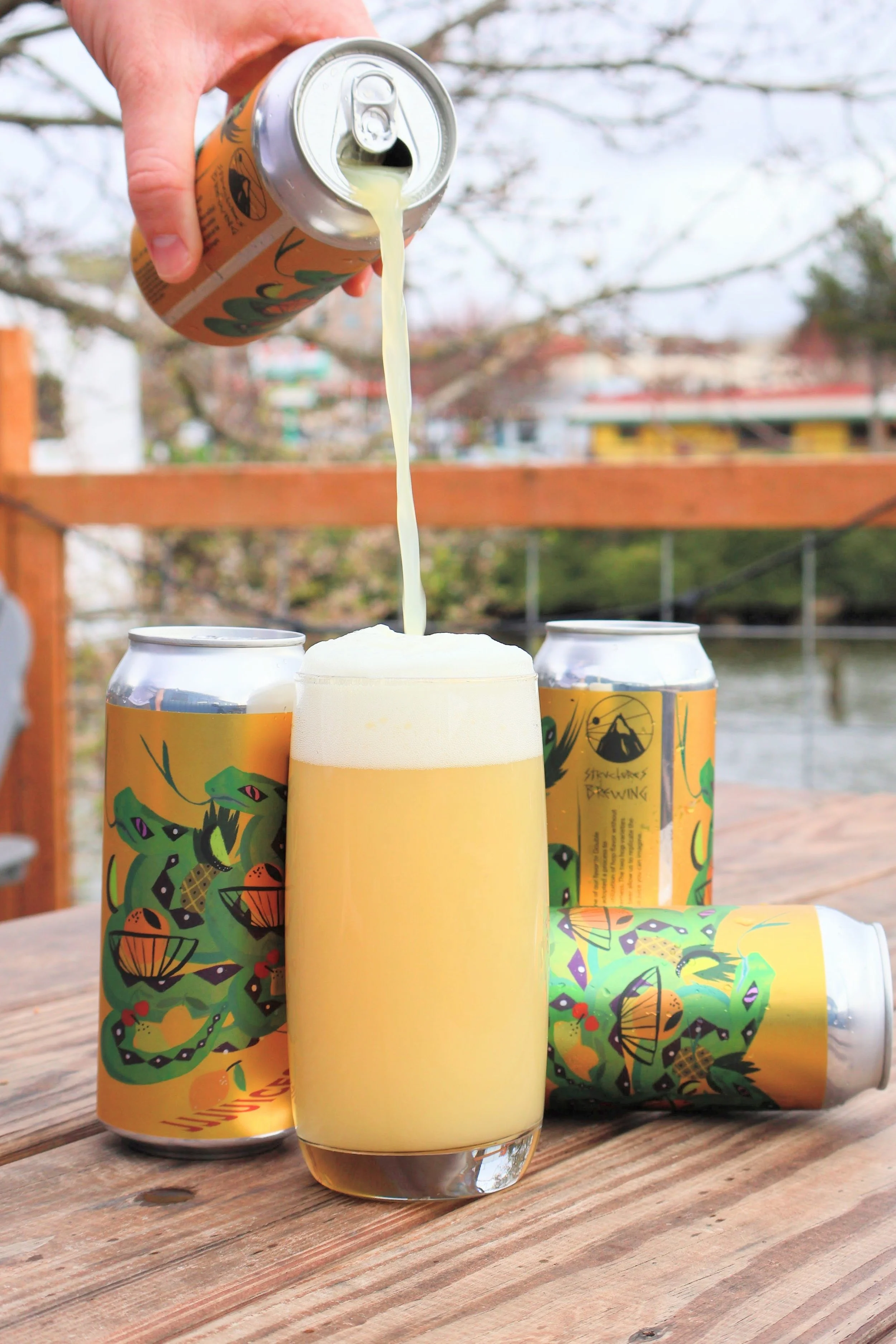A hand pouring a yellow beverage from a can into a glass, with three cans featuring colorful dragonfly and leaf designs on a wooden table outdoors, overlooking a river with trees and buildings in the background.