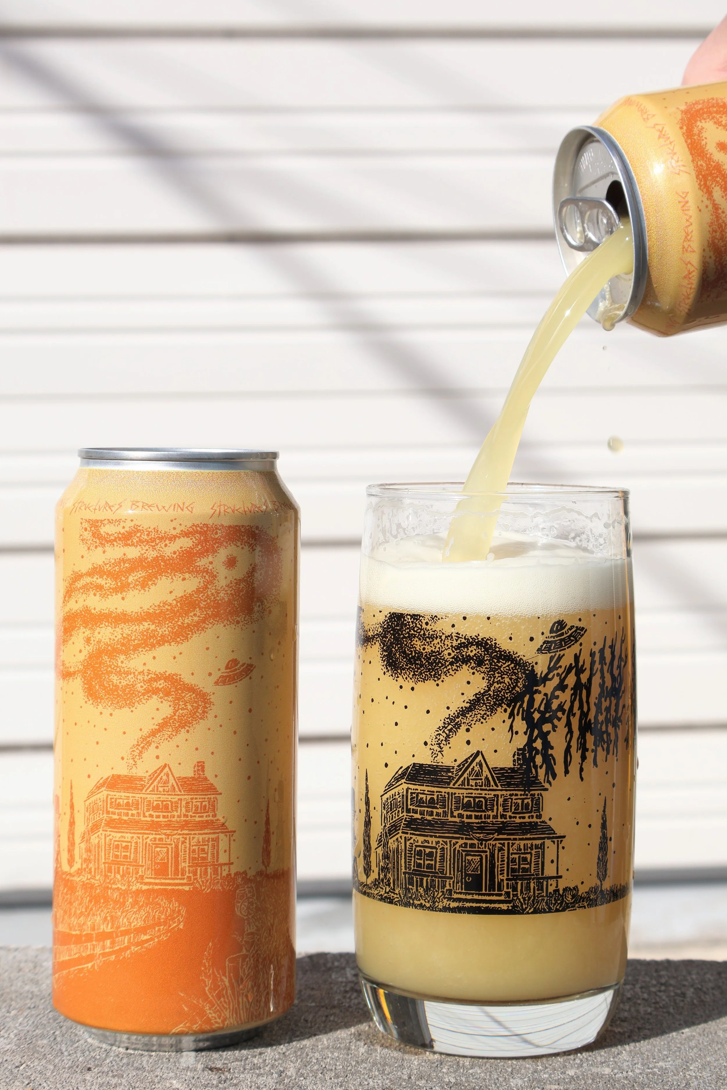 A can of beer being poured into a glass, with an identical can and a glass of beer on a concrete surface in front of a white slatted wall.