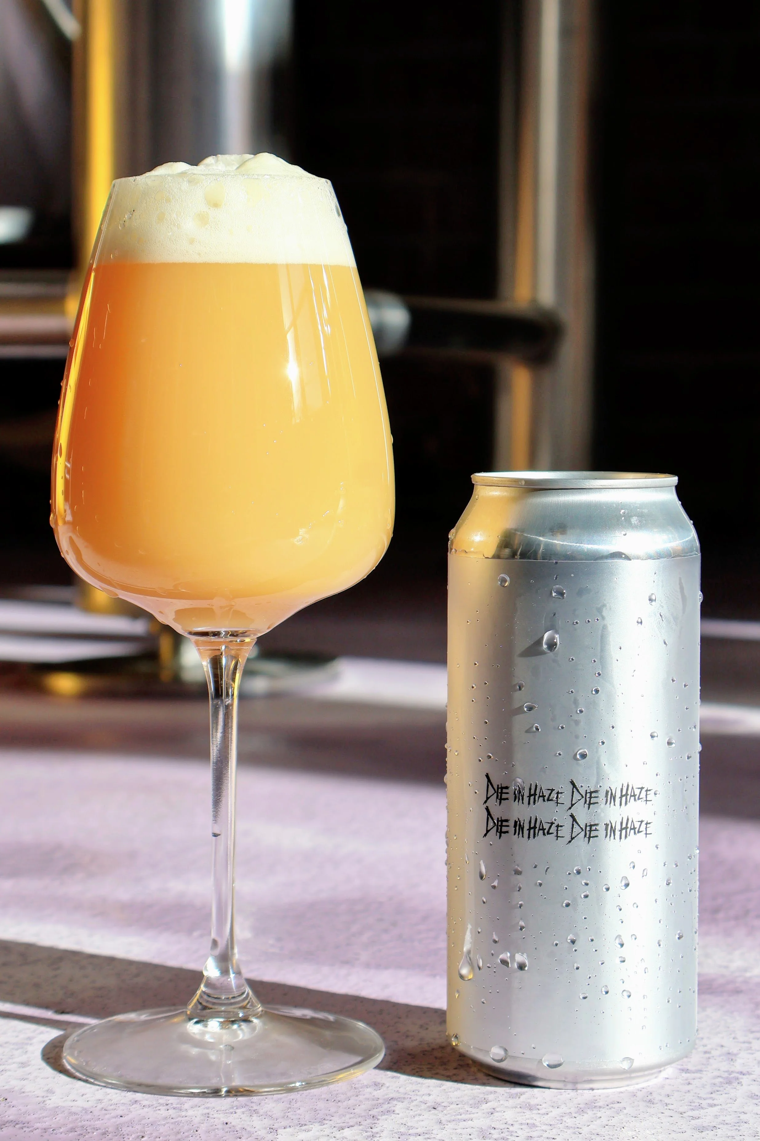 A can of beer with the words "Die In Haze" on it, beside a glass of hazy, pale beer with a foamy head.
