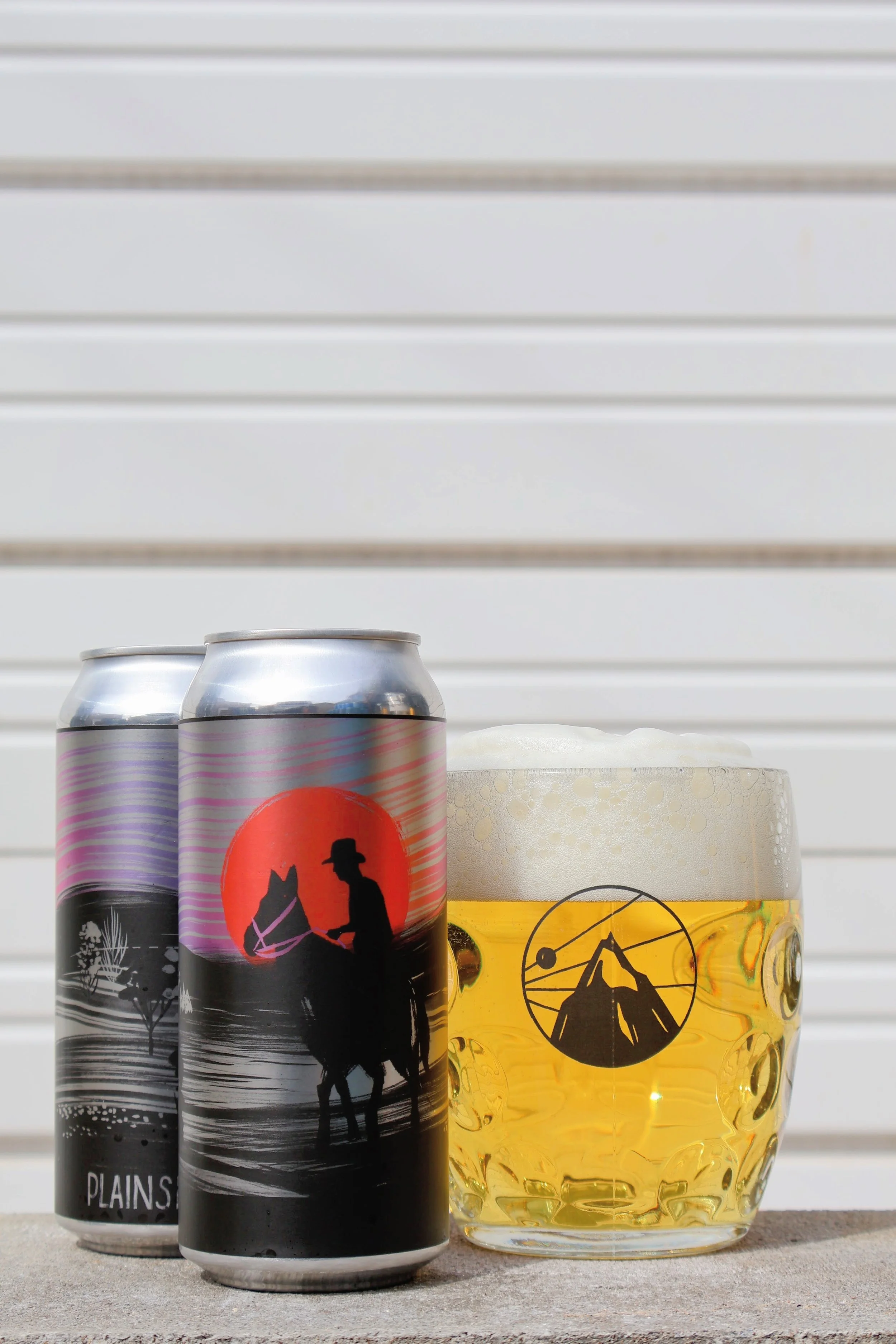 Two cans of craft beer and a glass of beer with a mountain and sun logo on a concrete surface with a white horizontal slat background.