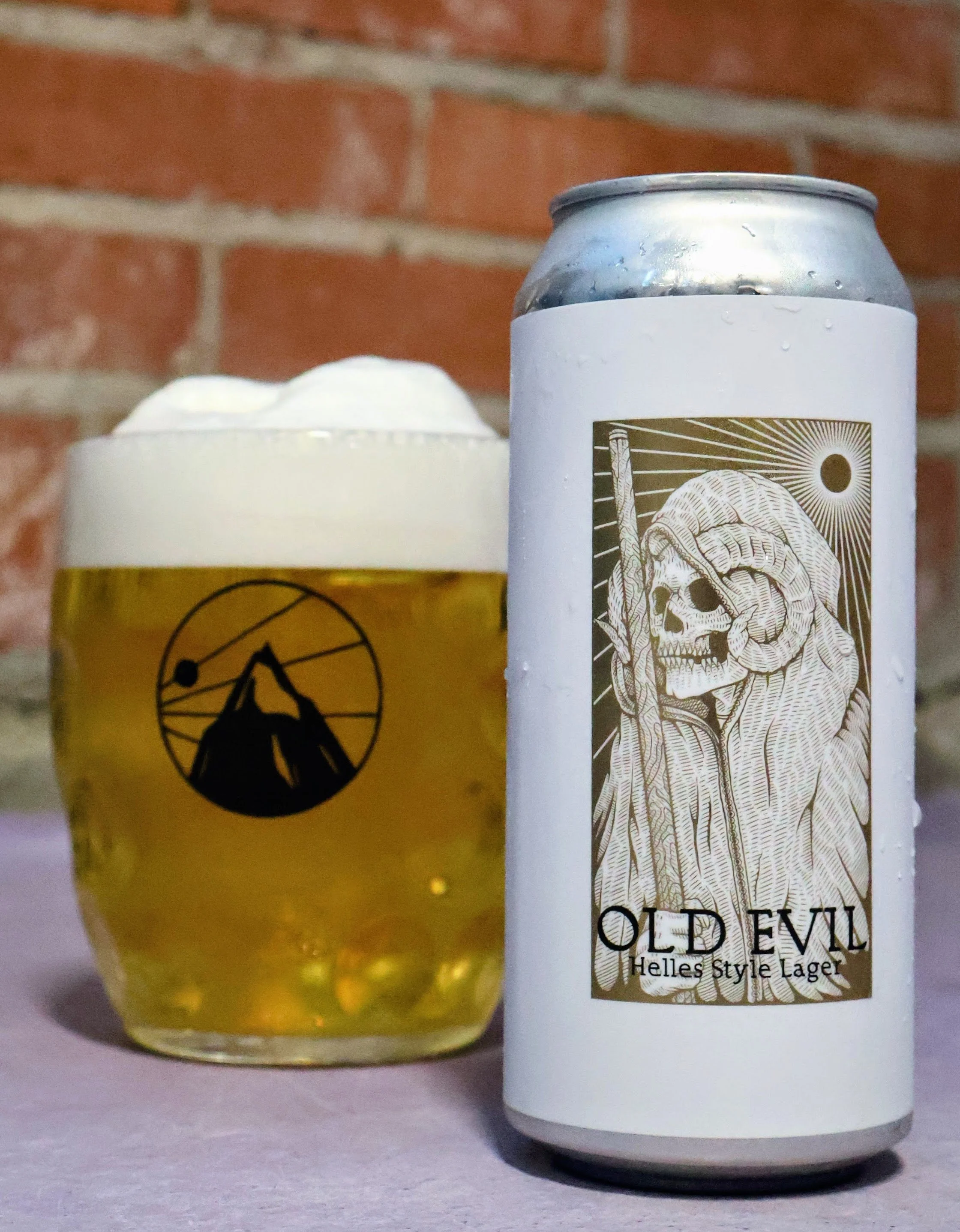 A can of Old Evil Helles Style Lager beer next to a glass of beer with foam on top, both placed on a surface against a brick wall.