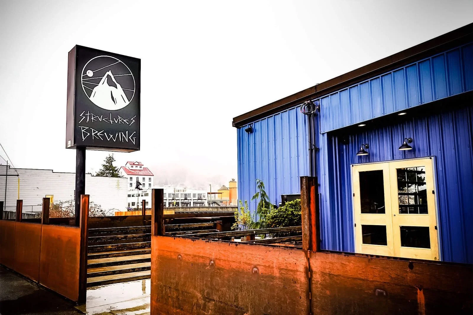 Street view of a blue building with yellow door, a street barrier, and a sign for Strukhoes Brewing with a mountain logo.