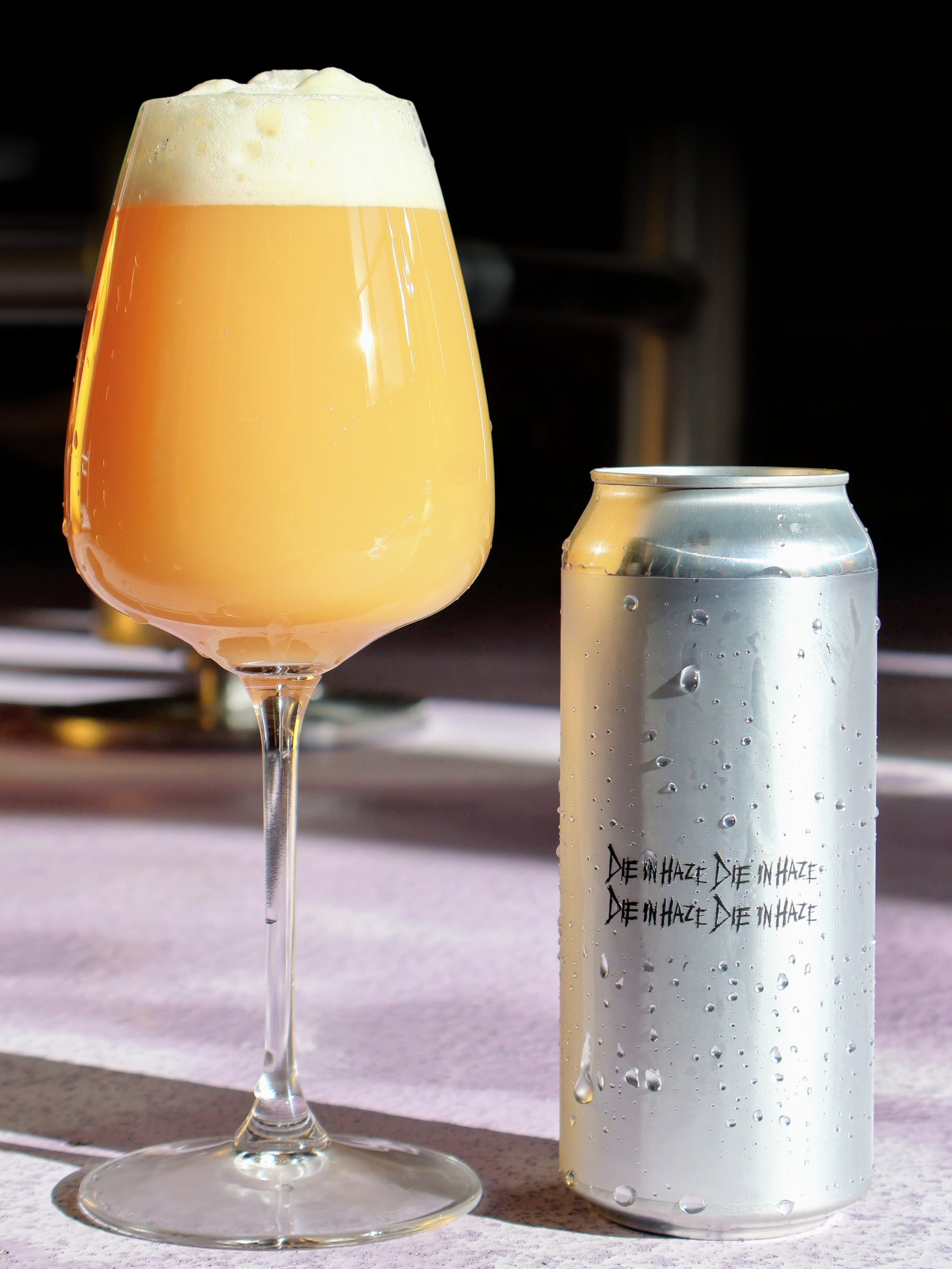 A glass of hazy, golden beer with a foamy head next to a silver beer can with condensation and the words "DIE IN HAZE" repeated on it.